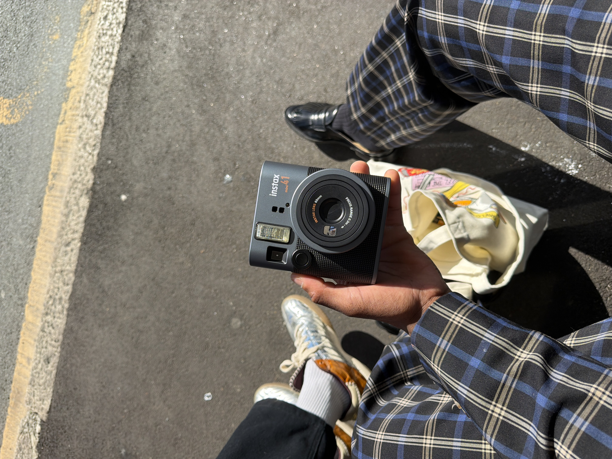 Captured from a high-angle perspective, the image features a person sitting or crouching outdoors on asphalt pavement with a textured white and yellow painted line, suggesting a street, path, or parking area. Bright sunlight illuminates the scene, casting distinct shadows and indicating a clear, sunny day.
The central focus is a person's hand holding a grey Fujifilm Instax mini 40 instant camera. Prominently displayed text on the camera includes "instax mini 40," "FUJIFILM," "INSTAX LENS 60mm," and "FOCUS RANGE 0.3m ~ ∞." The camera's lens is in the center, with the flash unit visible to its left.
The person is wearing plaid trousers with a pattern of black, blue, and light-colored lines. Their footwear varies: a dark, possibly leather loafer is visible on the left foot, while the right foot wears a shiny, metallic silver sneaker with white laces and a distinctive orange or brown trim, paired with white ribbed socks.
A cream-colored tote bag with colorful, indistinct graphic elements and some illegible text is partially visible beside the camera, resting on the ground or the person's lap. The setting is consistent with an outdoor location, potentially within the Folkestone and Hythe District.