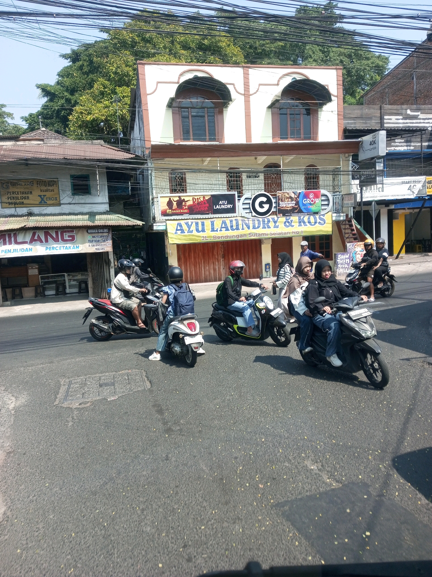 In the city of Malang, Indonesia, a sunny day unfolds on a street with a mixture of commercial buildings and trees. Several individuals are seen riding motorcycles. In the foreground, a person wearing a blue shirt and jeans rides a white scooter, while another individual in a black shirt and jeans, wearing a black headscarf, rides a dark-colored motorcycle with a passenger. Further back, a person in a white shirt and shorts rides a red and black motorcycle, and another person in a dark shirt and helmet is on a dark motorcycle with a passenger. A fourth motorcycle, also dark-colored, carries two people.
The buildings lining the street exhibit varying architectural styles. One prominent building has a cream-colored facade with arched windows and brown trim. Adjacent to it is a lower building with a corrugated metal roof and awnings. Signs for businesses like "AYU LAUNDRY" and "MILANG PERCETAKAN" are visible. The street itself is paved, with a manhole cover visible in the lower portion of the image. Overhead, a complex network of electrical wires crisscrosses the sky. The overall scene captures a typical street view in a bustling Indonesian city.