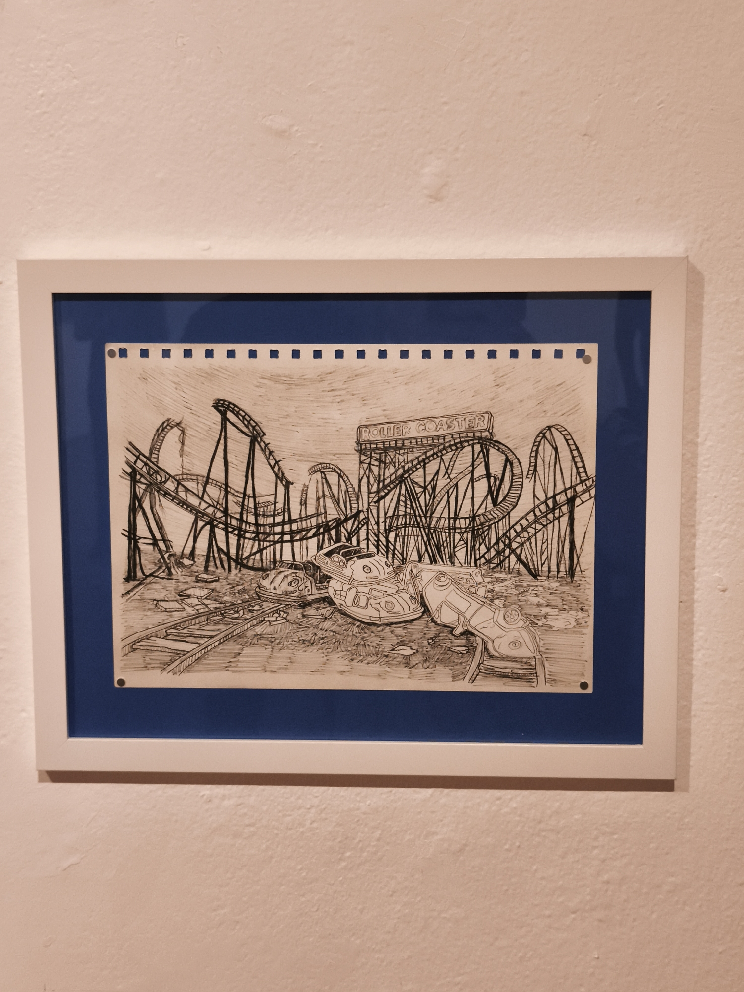 The image shows a framed black and white drawing of an abandoned amusement park with a roller coaster as a central element. The drawing is on paper with perforation at the top, suggesting it was torn from a notebook. The roller coaster structure, with the sign reading "ROLLER COASTER", is surrounded by the wreckage of cars and other park elements, giving the impression of ruin and decay. The drawing is mounted within a blue border and then placed inside a white frame, which is hanging on a white wall. The scene is not recognizable as a specific location in Querétaro, Mexico, as it appears to be a generic abandoned amusement park. There are no people visible in the scene.