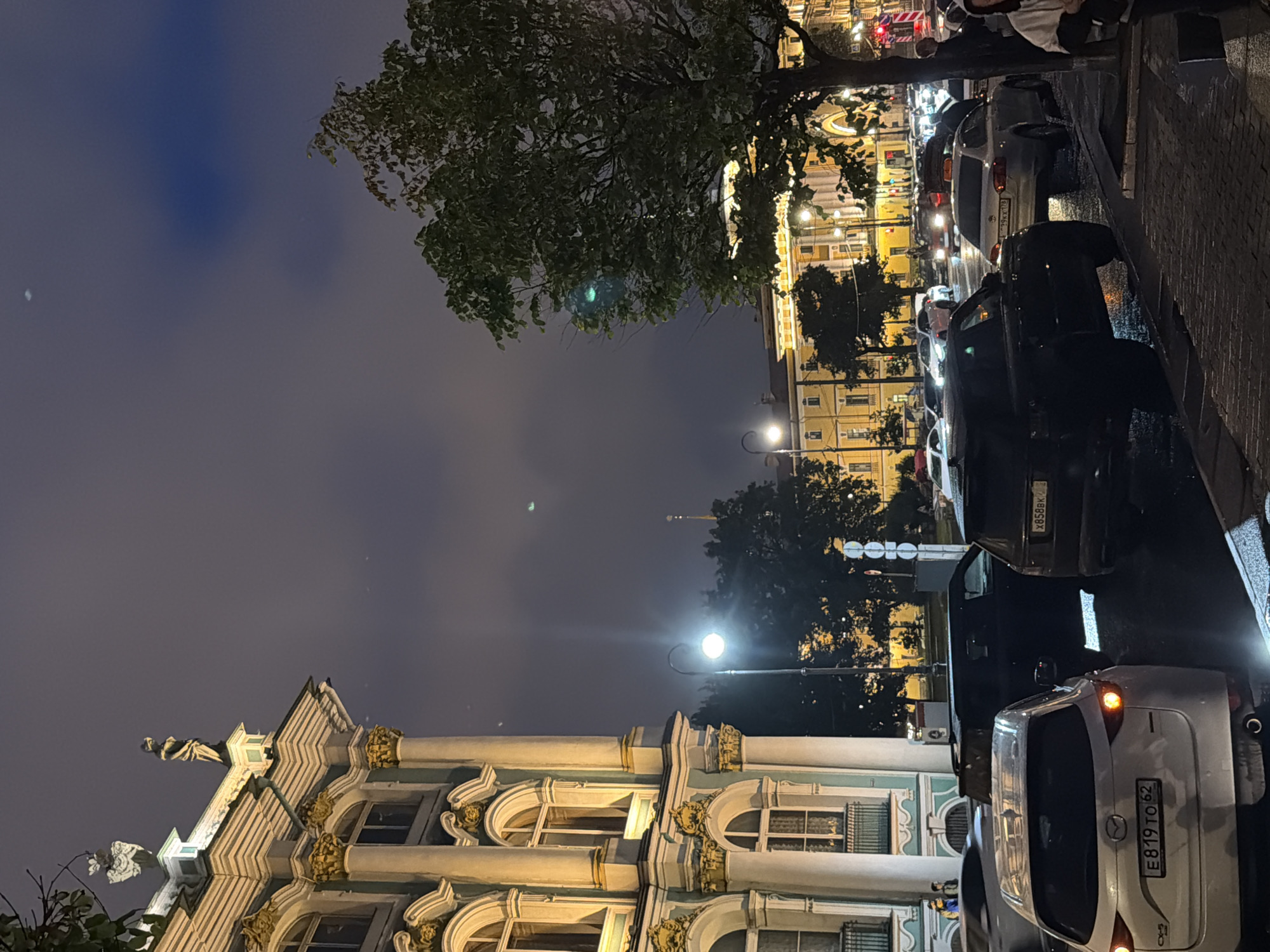 This is a night-time outdoor shot of a street scene in an urban environment. On the left, a grand building with ornate architecture, featuring classical columns, arched windows, and decorative elements, is illuminated by warm lighting. In front of the building and extending to the right, a street is lined with parked and moving vehicles, including SUVs, sedans, and a white Mazda CX-5 with a license plate that reads "E819 TO 62". A black SUV is parked prominently in the foreground. The street is wet, suggesting recent rain. A tall street lamp with a glowing bulb stands near the building. Trees with lush foliage are visible, some illuminated by streetlights and building lights. In the background, other buildings with illuminated windows and architectural details line the street, and a tall spire can be seen in the distance. The sky is dark and cloudy, with some faint lights visible in the upper sky. A few figures are visible near the parked cars and on the sidewalk.