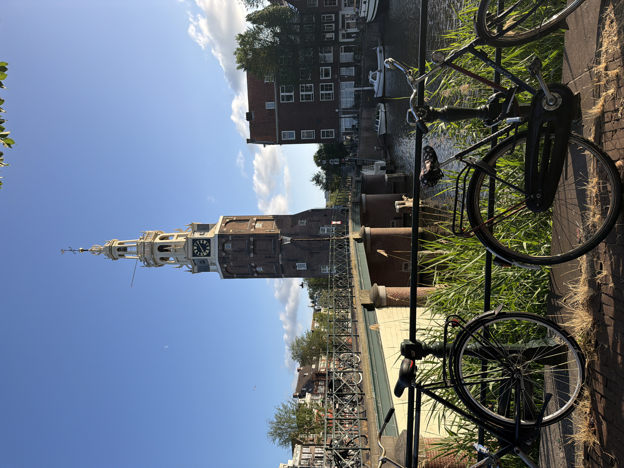 The image shows a picturesque view of a canal in Amsterdam, Netherlands, on a sunny day with a clear blue sky and scattered clouds. In the background, the iconic Montelbaanstoren tower stands tall, its clock face visible. A bridge spans the canal, with several bicycles parked along its railing. On the canal itself, small boats are docked, and the water reflects the surrounding buildings and the sky. In the foreground, two bicycles are positioned in front of a railing, with tall grasses growing behind them. The scene depicts a typical Amsterdam canal landscape, characterized by its historic architecture, waterways, and bicycles.