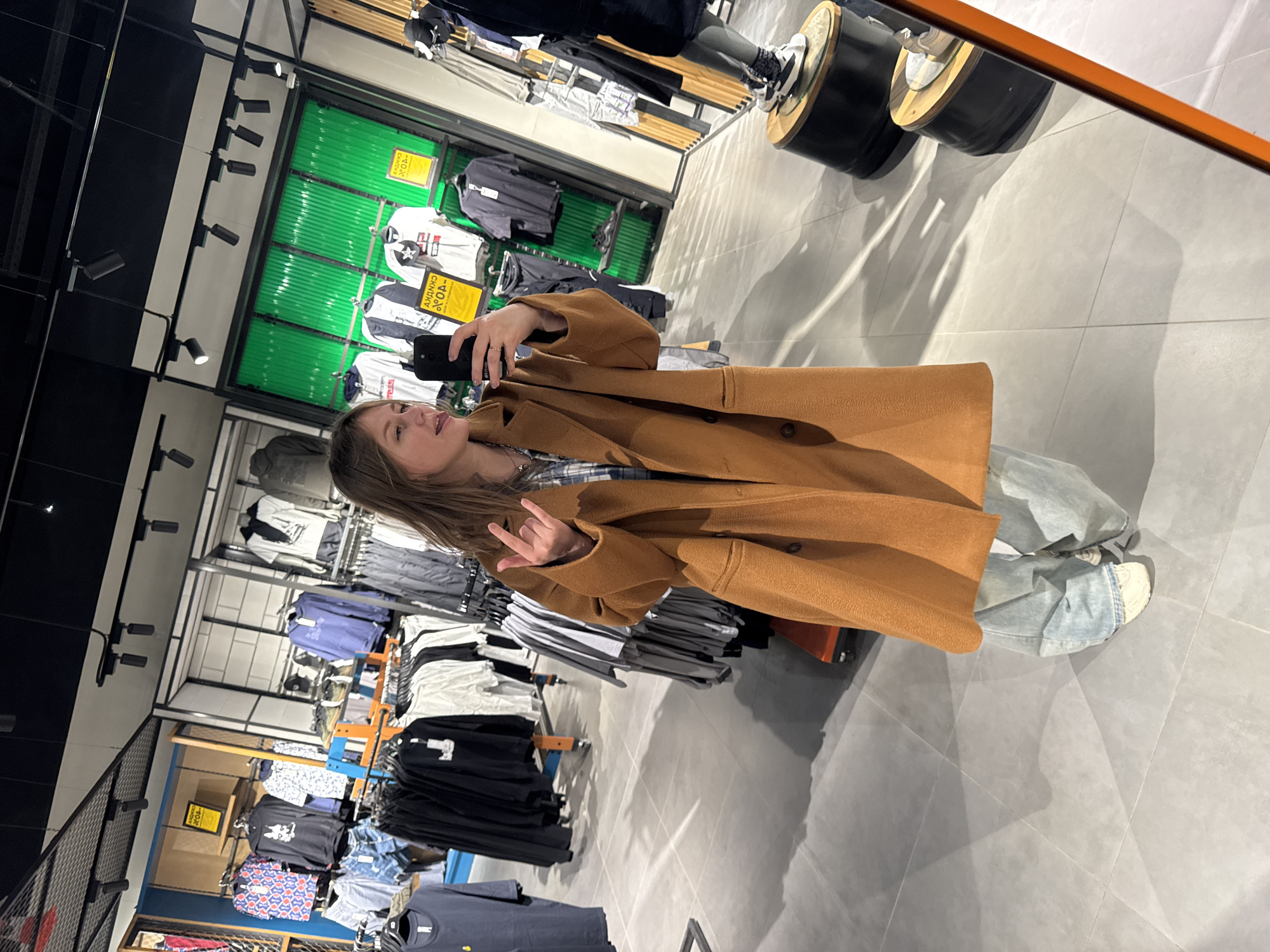 A young woman is depicted taking a mirror selfie inside a clothing store, her image appearing as a reflection. She is seen from the chest down to her feet, holding a dark smartphone in her right hand. Her left hand is raised, performing a "rock on" hand gesture. She has long, light brown hair and is wearing a camel-brown, long-sleeved coat over a blue and white plaid shirt, light-wash blue jeans, and white sneakers.
The setting is a retail environment, characterized by numerous clothing racks displaying various garments, including t-shirts, shirts, and trousers. A prominent feature in the background is a green slatted wall section, used for merchandising. Overhead, black track lighting fixtures illuminate the store. The floor is a light gray tiled surface. In the upper right quadrant of the image, the reflection of a cylindrical display stand with a mannequin's lower body wearing sneakers is visible.
Several yellow signs with text are visible on the clothing displays. The most prominent one clearly shows "АКЦИЯ -40%", which translates from Russian as "PROMOTION -40%", indicating a sale. The overall scene suggests a casual shopping experience during daylight hours in a well-lit, modern store, consistent with the provided location context of Perm, Russia.