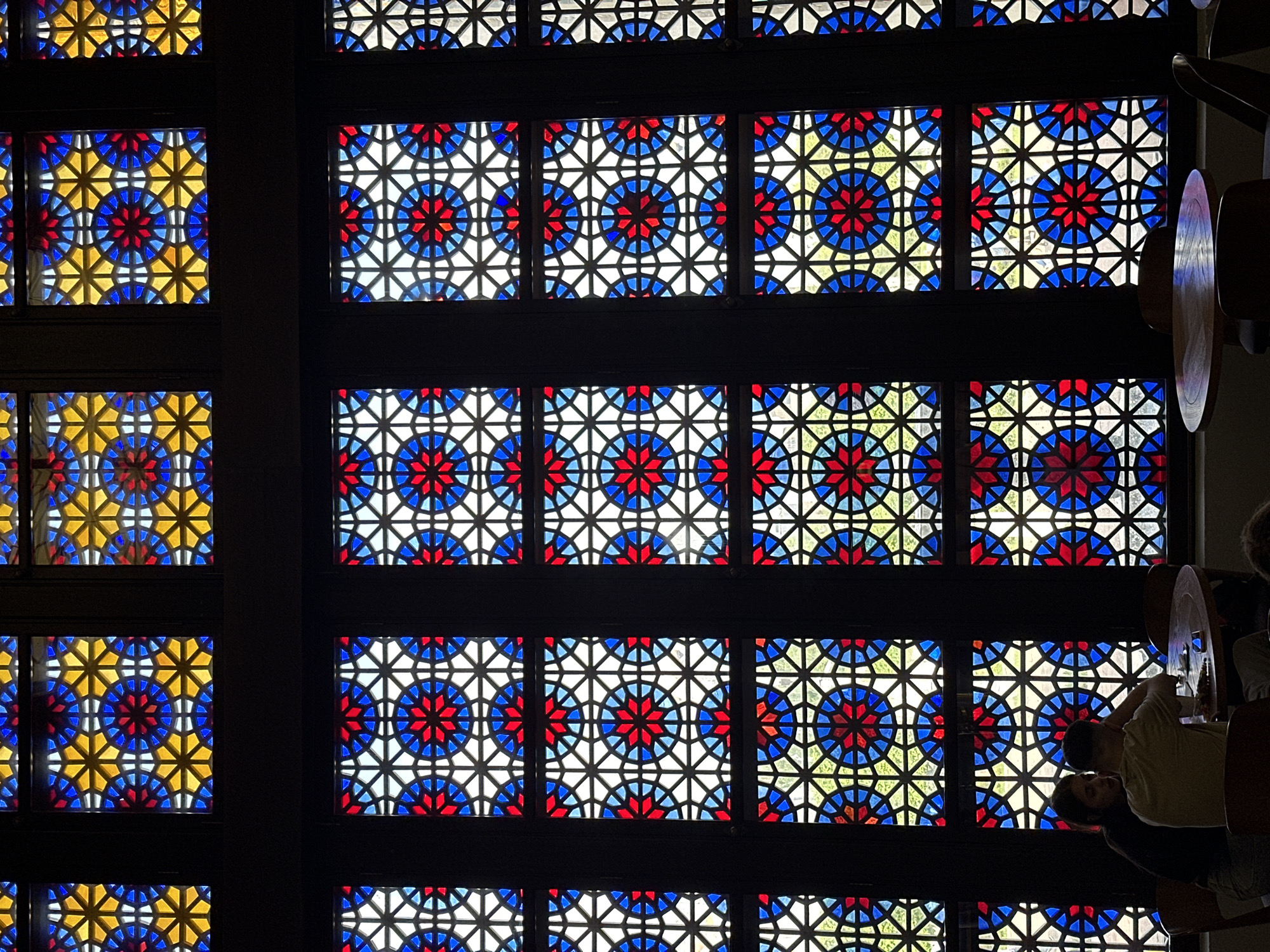 Captured from a high-angle perspective, the image showcases an interior featuring stained glass windows. The glasswork exhibits a repetitive pattern of circular designs with alternating red, blue, white, and yellow hues. The windows are divided by dark frames. The right side of the image features portions of two people, one wearing a light-colored shirt, with part of a wooden structure visible beside them. The photograph was taken in Unknown, Unknown.