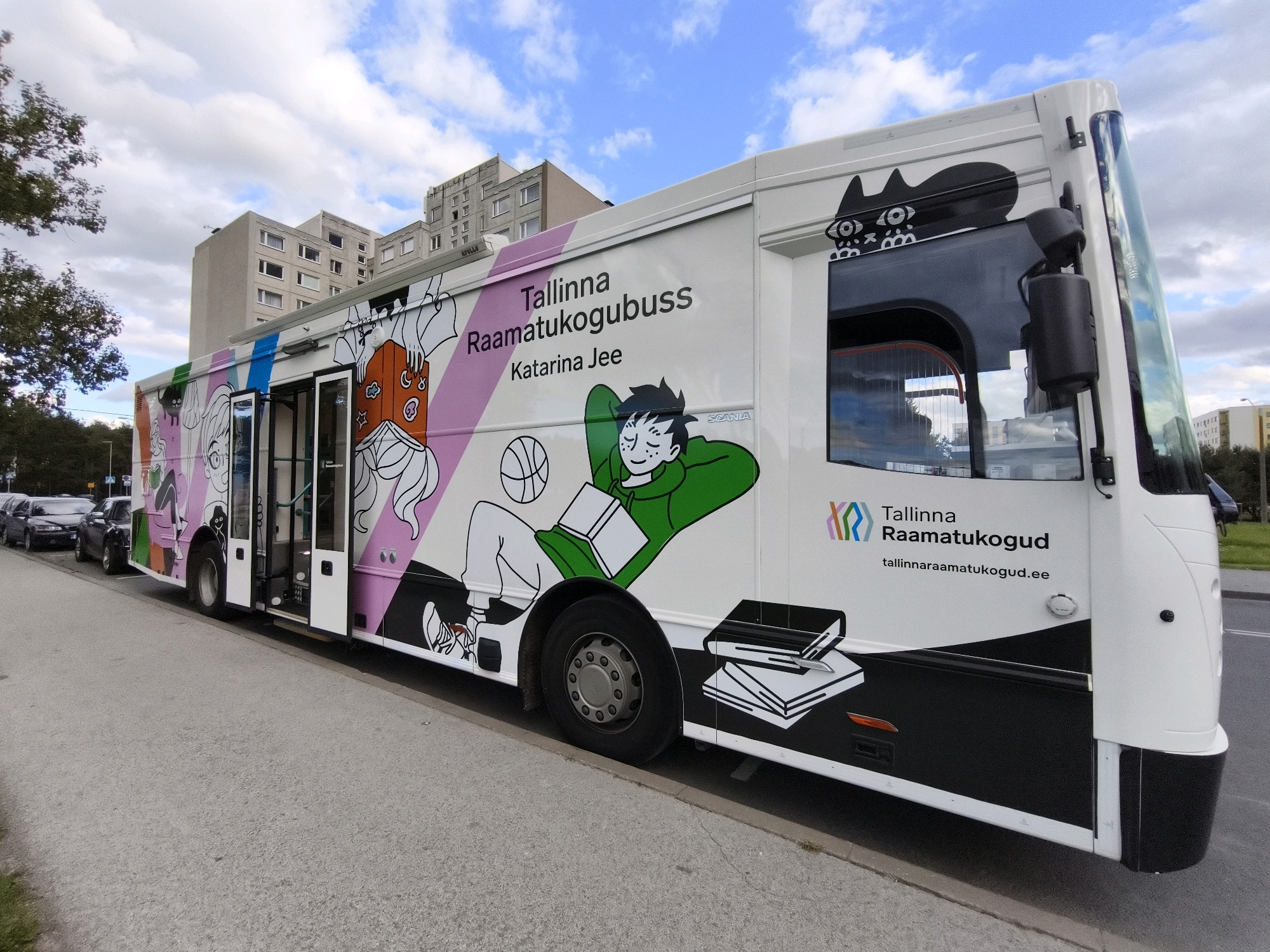 A white bus with colorful cartoon illustrations is parked on the side of a street in Tallinn, Estonia. The bus is decorated with images of characters reading, playing basketball, and holding books, along with the text "Tallinna Raamatukogubuss Katarina Jee" and the website address "tallinnaraamatukogud.ee." The bus is a mobile library, or "raamatukogubuss," a service provided by the Tallinn Central Library. In the background, apartment buildings and trees line the street. The sky is partly cloudy. Several cars are parked in front of the bus.