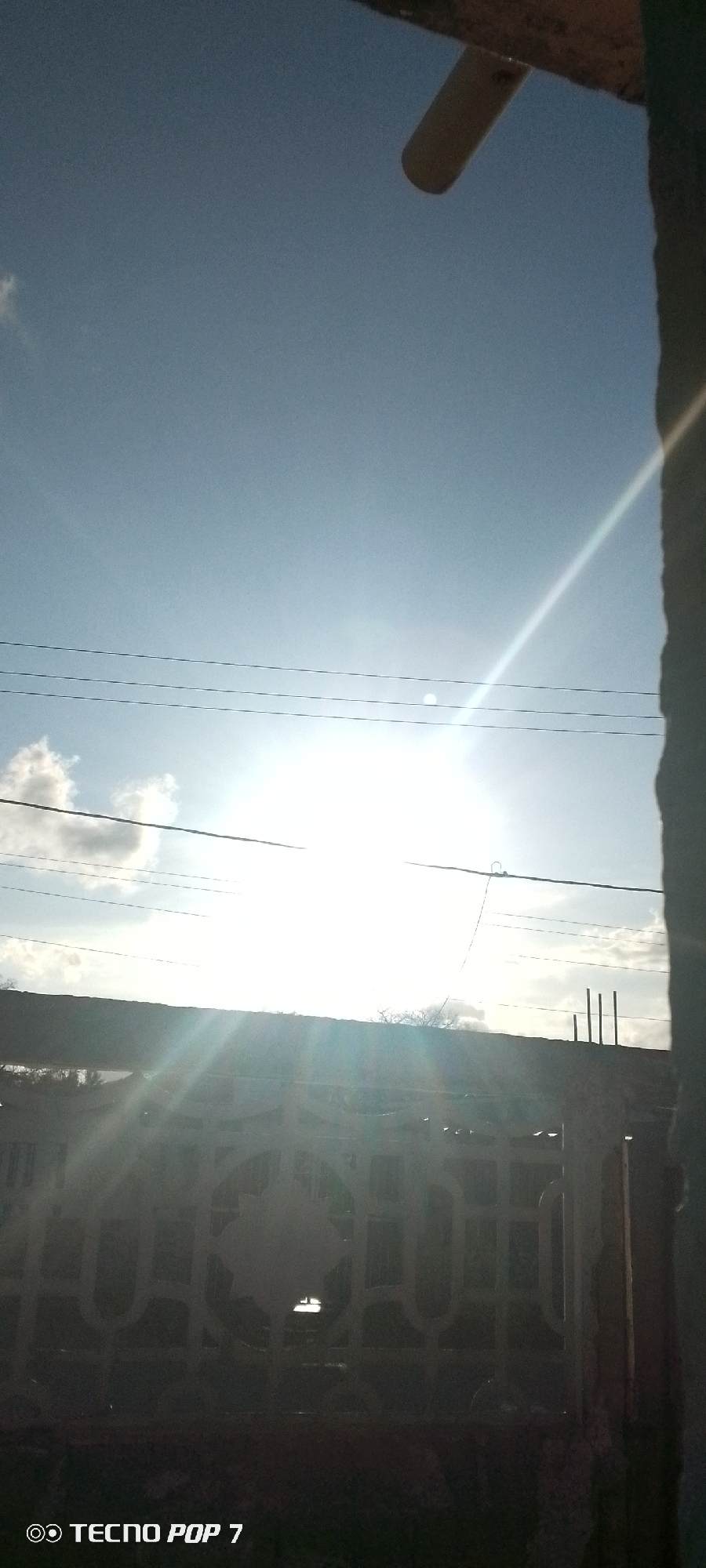 The image shows a view of the sky through a gated fence, possibly in Altagracia, Venezuela. The sky is bright blue with some clouds visible near the horizon. The sun is high in the sky, creating a strong glare. Power lines run horizontally across the center of the frame. The gate is made of dark metal, partially obscuring the view. A wall or building is visible on the right side of the image, slightly out of focus. The bottom of the image has the inscription "TECNO POP 7".