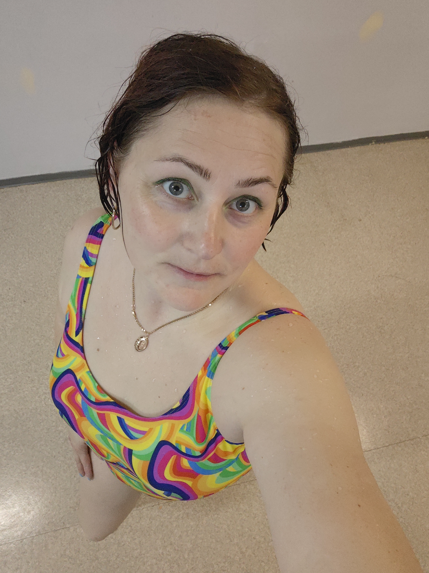 A woman with wet, dark hair, light skin, and blue eyes, wearing a colorful patterned swimsuit and a gold necklace with a round pendant, is looking directly at the camera. She has what appears to be green eyeliner on her upper eyelids. Water droplets are visible on her skin and swimsuit. She is indoors, standing on a tan, tiled floor in front of a white wall. The location is Markovskii, Russia.