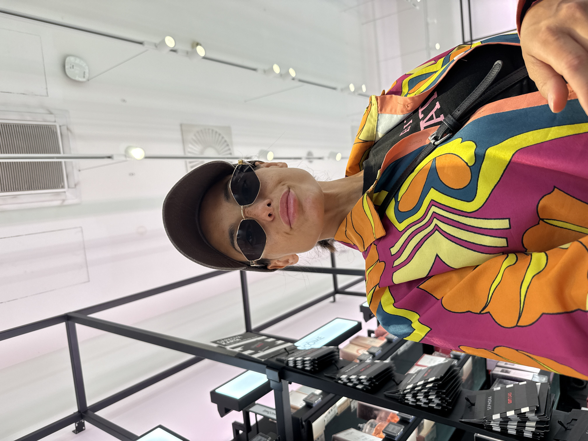 A woman wearing a baseball cap, sunglasses, and a colorful patterned jacket stands in a brightly lit store. She is looking towards the camera, her lips slightly pursed. Behind her, shelves are stocked with various beauty products and gift cards, some bearing the Sephora logo. The ceiling features overhead lighting and ventilation. The image appears to have been taken indoors, possibly in a makeup or cosmetics store.