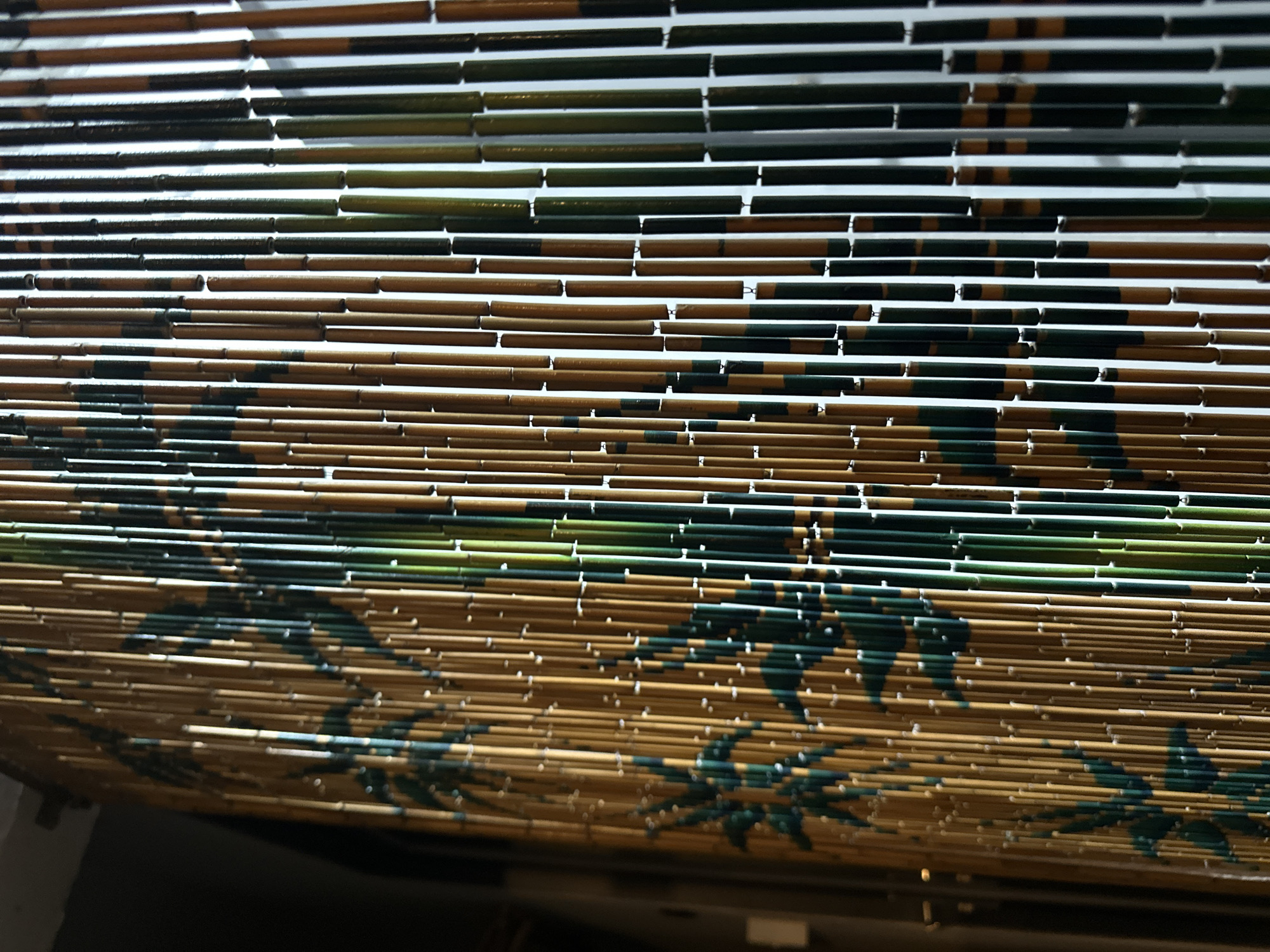 This is a close-up, eye-level, indoor shot of a beaded curtain made of bamboo sticks. The curtain is hanging vertically, and the sticks are of varying colors, predominantly natural bamboo brown and dark green. There is a repeating pattern of stylized green bamboo leaves painted onto some of the brown sticks. The lighting is somewhat dim, with the right side of the curtain appearing brighter due to an unseen light source, creating highlights on the bamboo. The background on the far left is dark and out of focus. There are no people or discernible actions depicted. The scene appears to be within a building, possibly a doorway or a room divider. The origin of this image is not specified, and therefore, it cannot be definitively placed within the city of Thủ Đức, Vietnam.