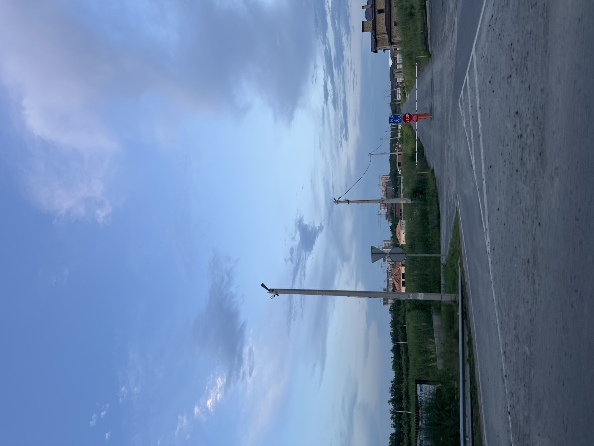 The image depicts a street scene in Yantarniy, Russia, during the daytime. The sky is filled with a mix of blue sky and scattered clouds, suggesting partly cloudy weather. In the foreground, a paved road with white markings is visible. To the left, a metal guardrail runs along the edge of the road, bordering a grassy area. A tall, slender lamppost stands prominently on the left side of the road.
Further down the road, a stop sign is positioned in the center, accompanied by a blue traffic sign indicating pedestrian and vehicle crossing. A white horizontal barrier, likely for traffic control, is extended across the road in front of the stop sign. On the right side of the road, a brick house with a brown roof is visible. In the background, construction cranes and unfinished buildings suggest ongoing development in the area, with a treeline forming the distant horizon. The overall environment appears to be a developing residential or suburban area. There are no people or animals visible in the image.