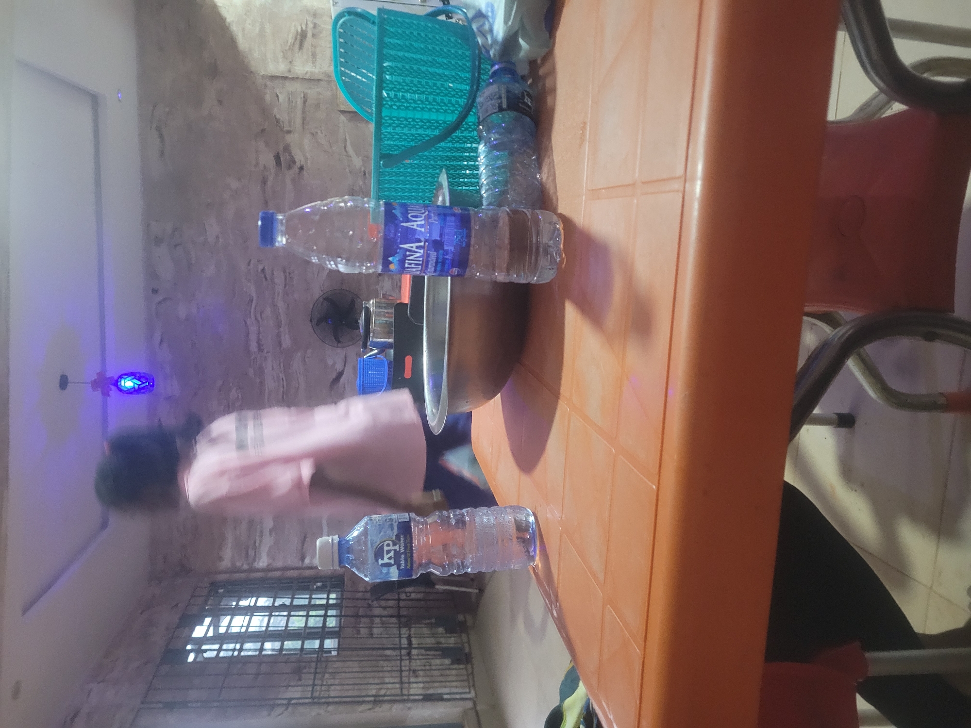 The scene appears to be indoors in Ogbunike, Nigeria, featuring an orange plastic table with several bottles of water (brands: 'KP Table Water' and 'Afinia Aqua') placed on it. A metal pot rests on the table next to one of the water bottles. A person wearing a light pink shirt and dark pants can be seen in the background. Other items include a blue plastic basket, and a fan mounted on the wall. The room appears to be lit by fluorescent lighting, including a purple decorative light fixture. The background shows a wall with a textured or worn appearance and a barred window. Part of a red and silver chair can also be seen on the right side of the table.