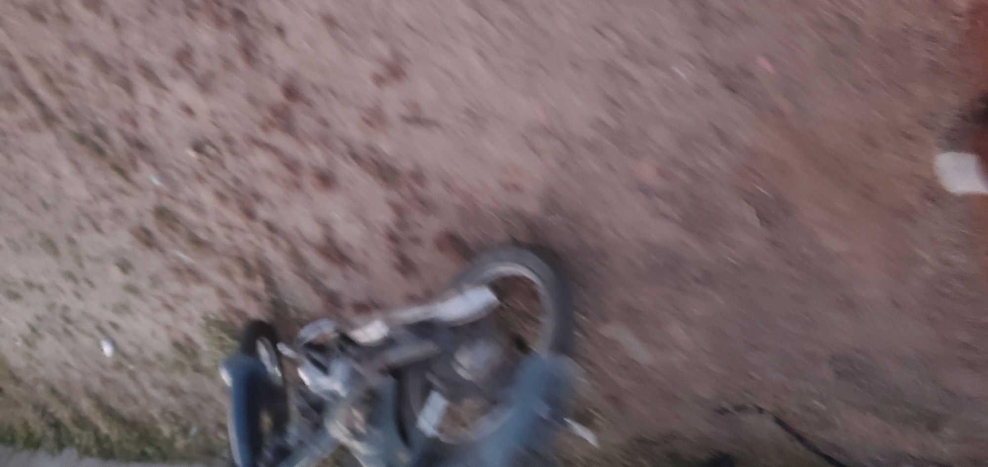 The image depicts a motorcycle lying on its side on a dirt surface in Numan, Nigeria. The motorcycle is partially obscured by blur, but appears to be a dirt bike or similar off-road model, with a dark-colored seat and silver-colored frame and engine components. The wheels are visible, with the rear wheel appearing to be slightly larger than the front. The surrounding environment is a sandy or dirt ground with sparse vegetation, suggesting an outdoor, possibly unpaved area. The lighting suggests it is daytime.