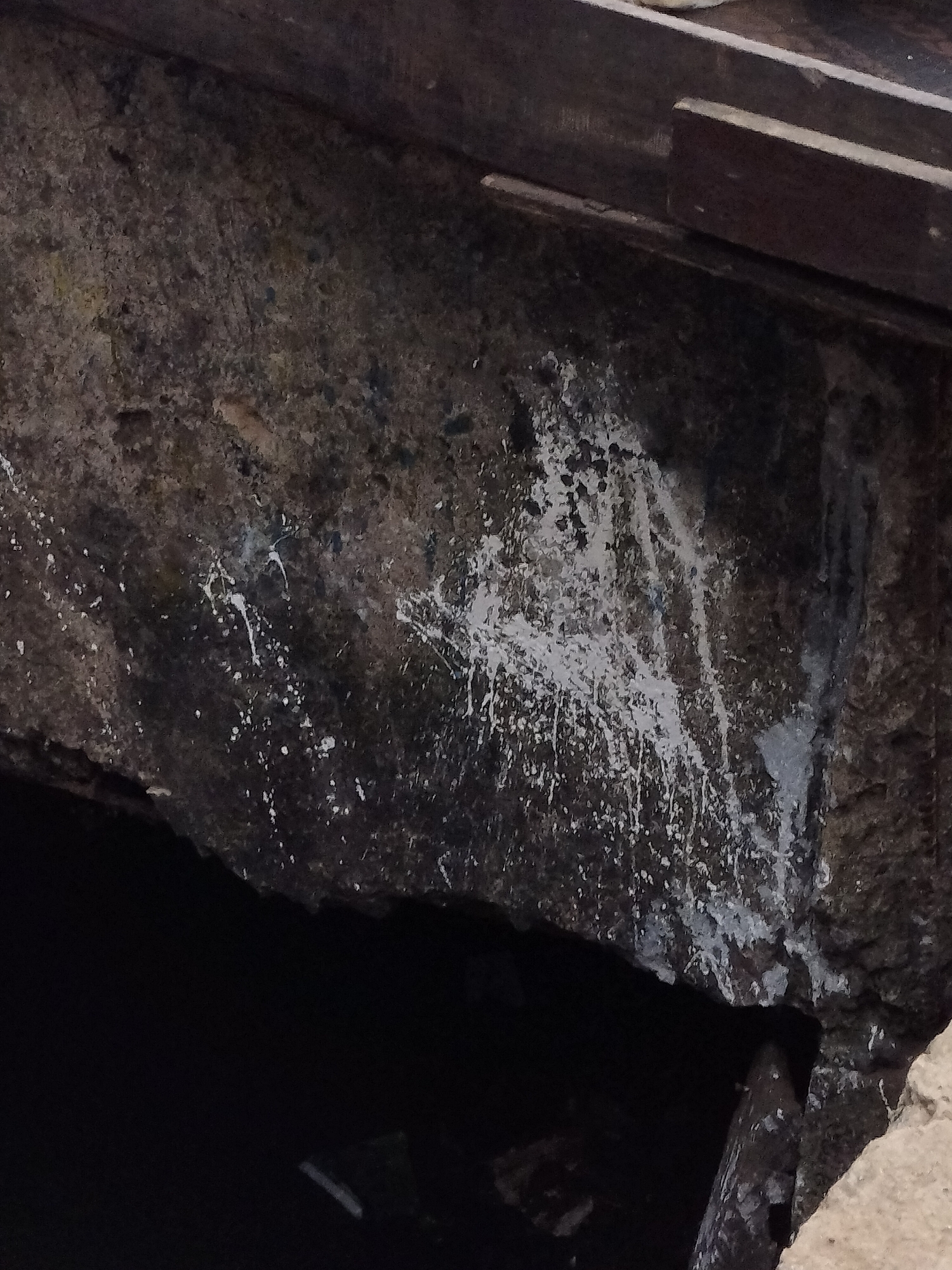 A dark, rough-hewn stone structure is visible, possibly part of a larger building or wall. Splatters of white paint are prominent on the stone surface, obscuring the underlying texture. A dark, possibly water-filled opening is located beneath the stone structure. The upper portion of the image includes a section of dark wooden beams. Without additional context, it is impossible to determine the location in Lagos, Nigeria, or other information.