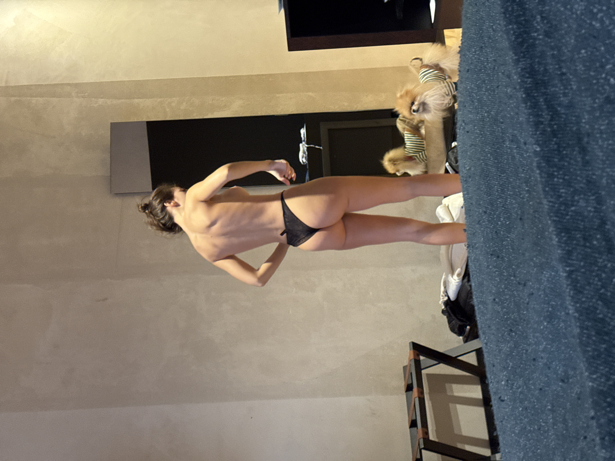 An adult female stands with her back mostly towards the viewer, slightly off-center to the right, in an interior room in Unknown, Unknown. She has an athletic physique with visible musculature in her upper back and arms, and her dark hair is gathered in a bun on top of her head. She is wearing only a black string thong, with the rest of her body bare. Her left hand is near her lower abdomen, and her right arm is bent at the elbow.
The room features light beige, textured walls. To the far left, a black metal luggage rack with brown straps is visible. A tall, dark-framed mirror stands vertically on the right side of the room, reflecting part of the wall and an obscured dark object. The lower foreground is covered by a textured blue/teal fabric, likely a bedspread.
Two small, fluffy dogs, resembling Pomeranians with light brown fur, are present on the floor in the mid-ground. Both dogs are wearing horizontally striped shirts (green and beige/brown). One dog is positioned closer to the viewer, to the right of the woman, looking forward. The second dog is further back, partially visible and reflected in the mirror. The scene is lit by dim, warm artificial light.