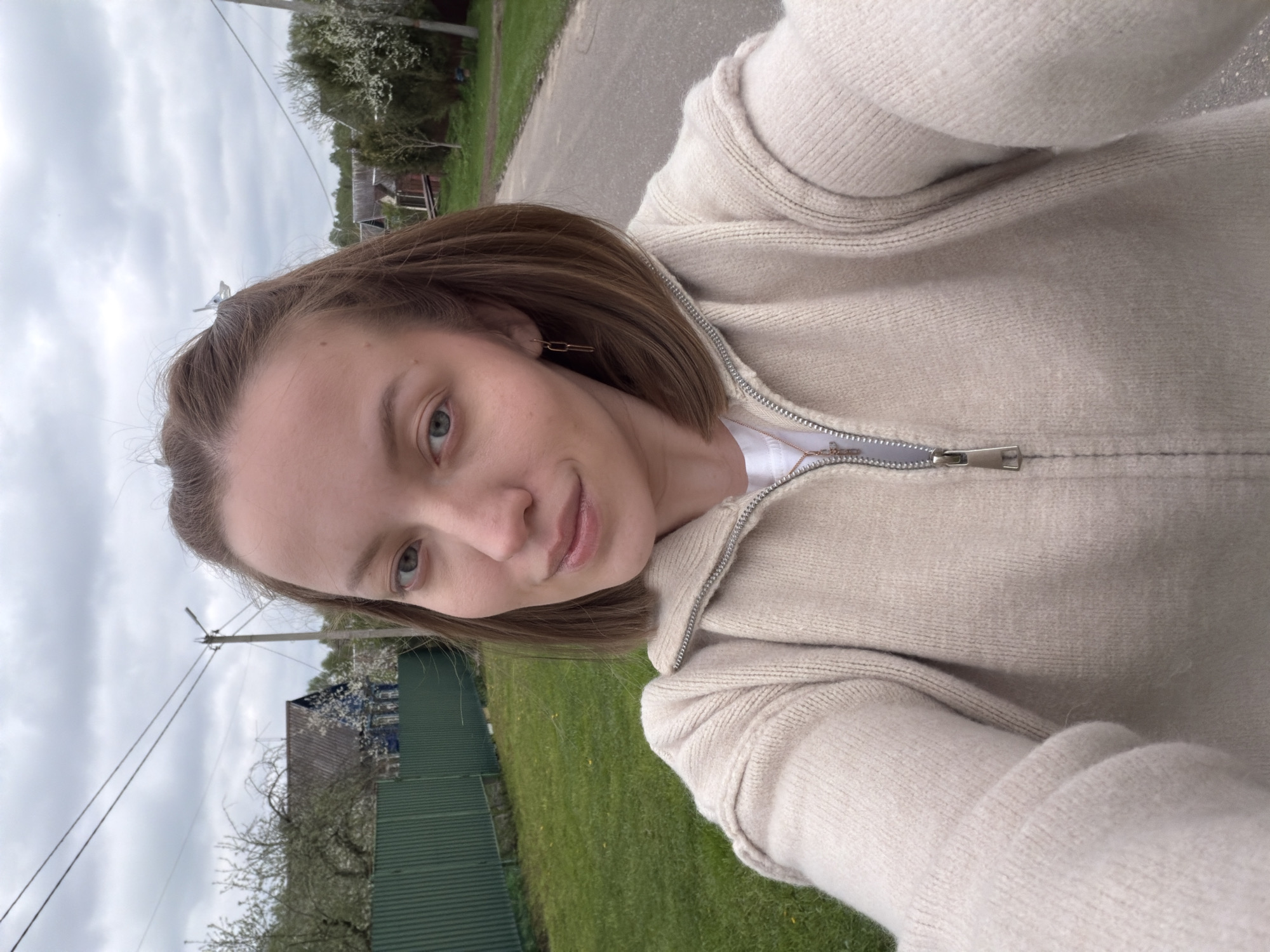 A woman stands on the side of a paved road in Reshetnikovo, Russia. She wears a beige knitted zip-up sweater over a white shirt and a thin gold chain. She has brown hair and fair skin. Behind her, a green lawn borders a dark green fence. A small house with a dark roof sits behind the fence, surrounded by flowering trees. Power lines run along the road. The sky is overcast.