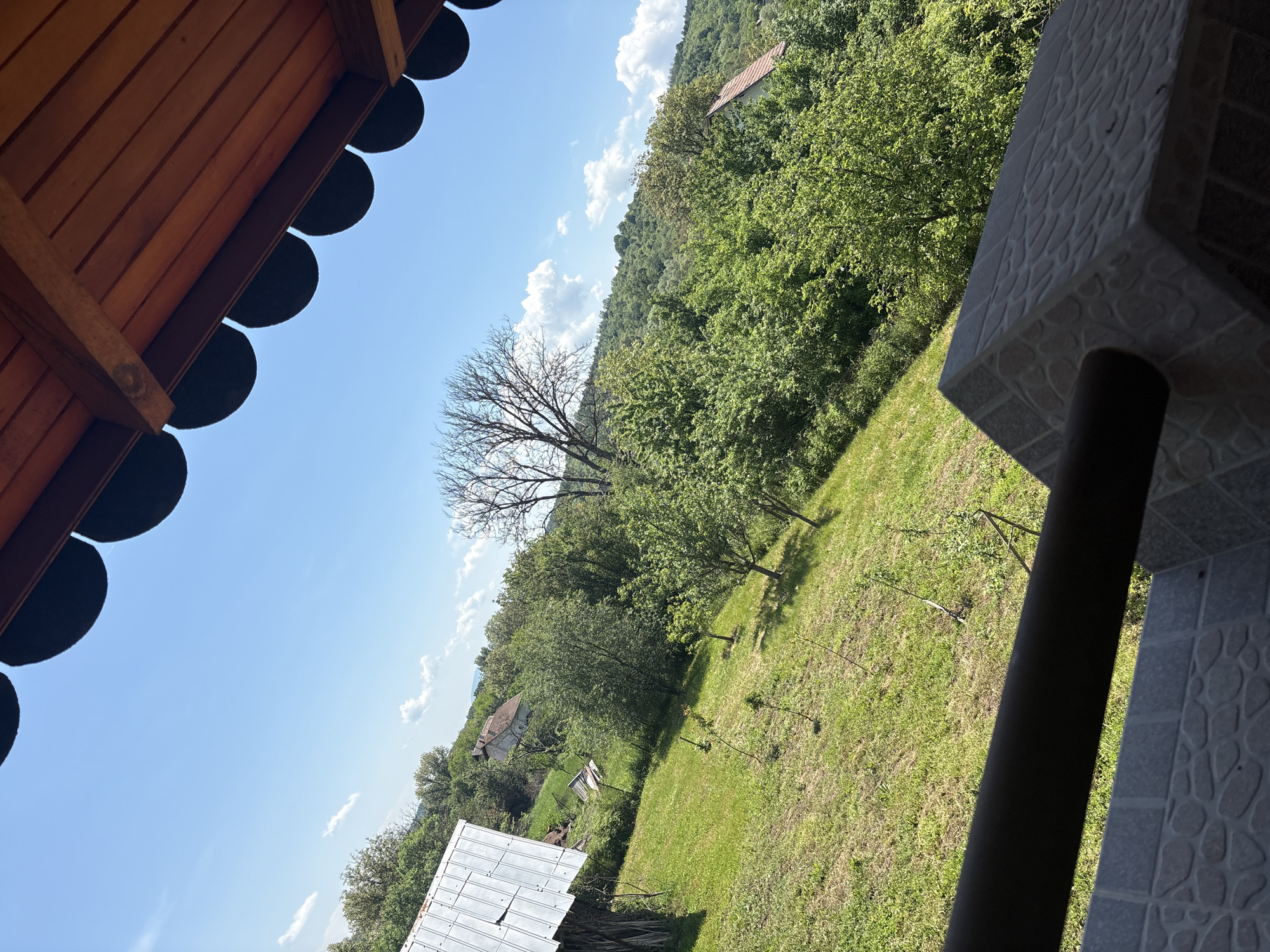 This media file presents a panoramic view of a rural landscape, likely captured from a raised vantage point such as a balcony or porch, with an unusual upward and sideways angle.
In the immediate foreground, to the upper left, is the underside of a wooden roof or overhang, featuring decorative scalloped edges and exposed wooden beams. To the right, a sturdy support structure is visible, made of textured grey tiles with an irregular pattern, alongside a dark, cylindrical pole or railing.
The central and lower portions of the image showcase a sloping grassy field or yard. Several trees are scattered across this area, including a prominent, tall deciduous tree completely devoid of leaves, contrasting with other trees that are lush with green foliage. Further into the midground, nestled among dense greenery, is a house with a reddish-brown roof, situated on a distant forested hillside that dominates the background. Another structure with a corrugated metal roof is partially visible in the lower-left.
The sky, filling a significant part of the frame, is a vibrant blue with scattered white, puffy cumulus clouds. The scene is brightly lit by natural sunlight, indicating a clear, sunny day, likely late morning or early afternoon, with discernible shadows on the grass and beneath the architectural elements. The mix of bare and leafy trees could suggest early spring or late autumn, or simply a dead tree within an otherwise verdant landscape. No people or visible text are present. The setting evokes a sense of tranquil, natural beauty in a countryside environment.