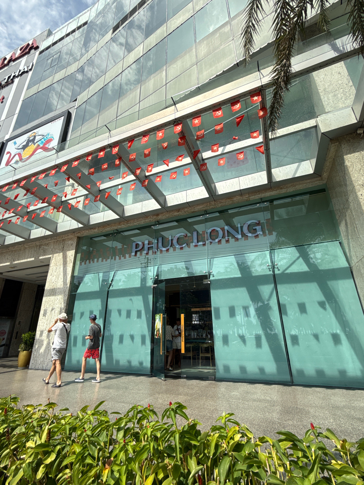 The image shows the entrance to a Phuc Long coffee shop located in Nha Trang, Vietnam. The facade of the building is made of glass and metal, with a large sign that reads "PHUC LONG" in white letters. A string of red flags with yellow stars and hammers and sickles is draped across the canopy above the entrance. In front of the entrance, two men are walking away from the camera. The man on the left is wearing a white t-shirt and grey shorts, and the man on the right is wearing a grey t-shirt and red shorts. In the foreground, lush green plants are in bloom. The sky is cloudy.