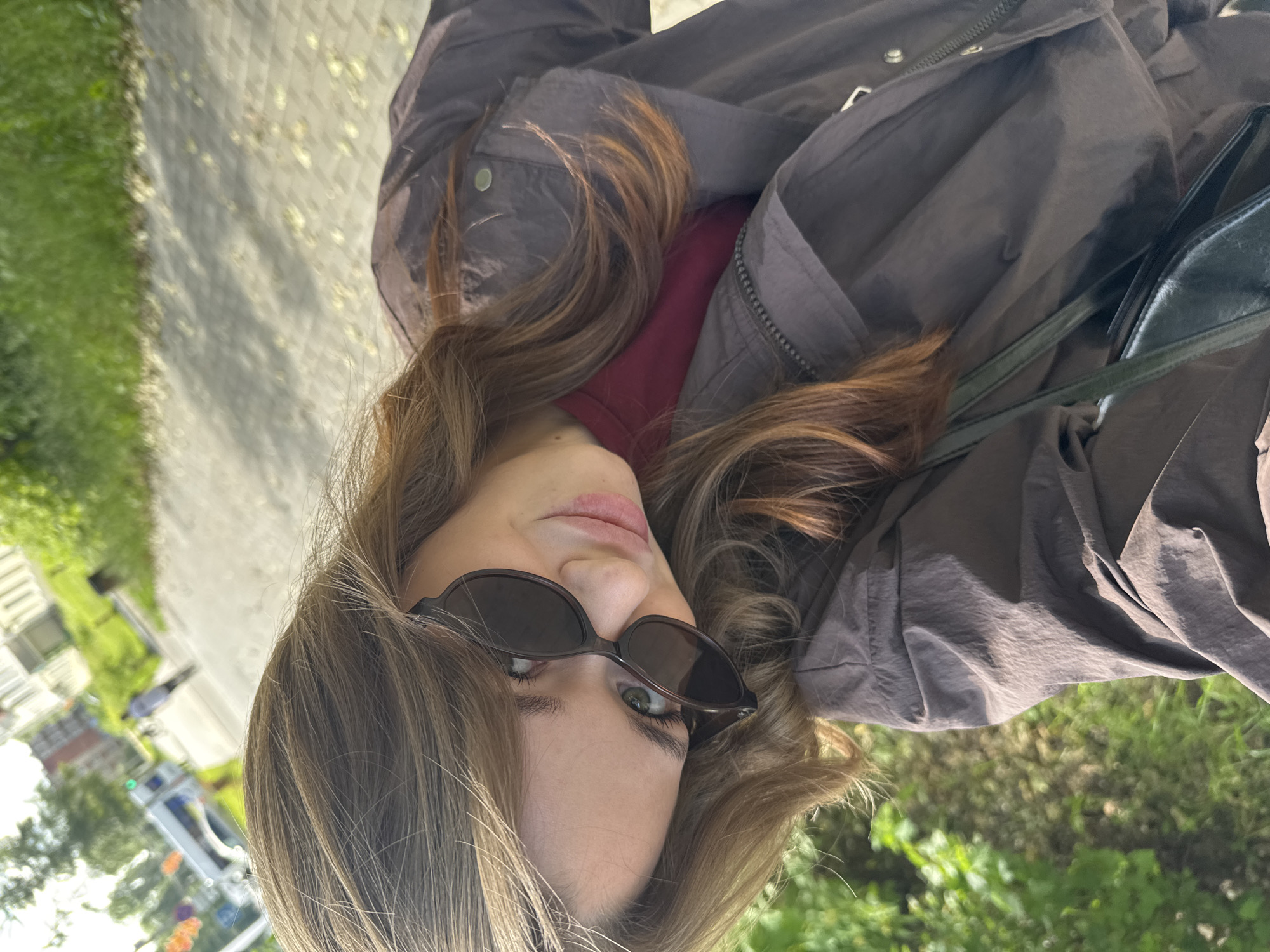 In the city of Chaikovsky, Russia, a young woman with long, wavy brown hair and green eyes poses for a selfie outdoors on a paved walkway. She is wearing dark sunglasses and a dark brown jacket over a maroon shirt. In the background, there is lush green foliage, a tree-lined street, and buildings. A white van and a pedestrian are visible further down the street. A dark bag is slung over her shoulder. The image is taken from a high angle, with the woman's face tilted towards the camera.