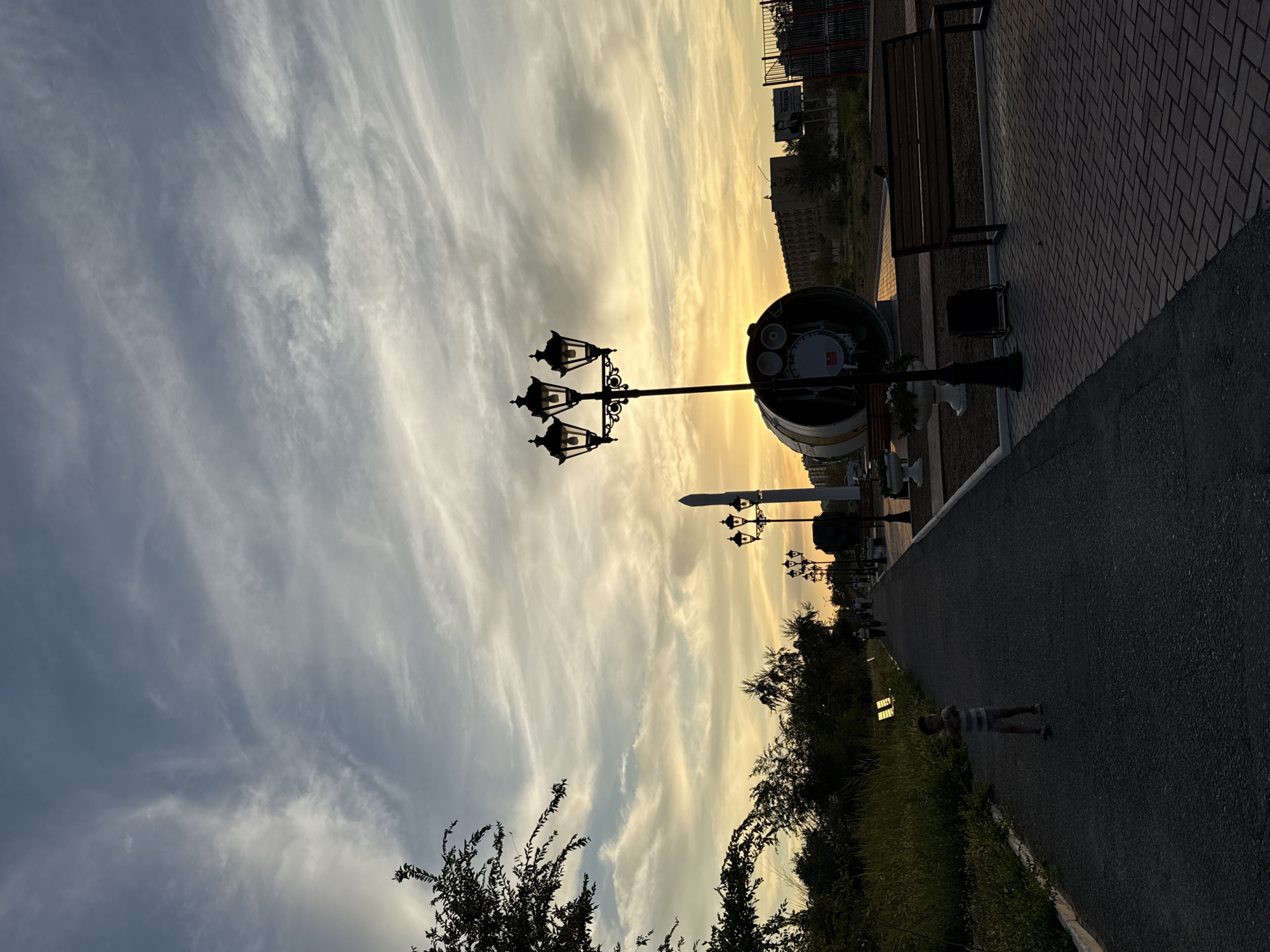 The image depicts a sunset scene in Baikonur, Kazakhstan, on a paved walkway lined with street lamps. A young boy in a striped shirt stands on the left side of the walkway, facing the camera. Further down the walkway, other people are visible, walking away from the camera. To the right, a large, cylindrical object, possibly a piece of rocket machinery, is displayed with a prominent street lamp in front of it. A wooden bench is situated next to the display. The sky is filled with wispy clouds, illuminated by the setting sun, casting a warm glow over the scene. Trees and grass border the walkway on the left, and buildings and a fence are visible in the background on the right.