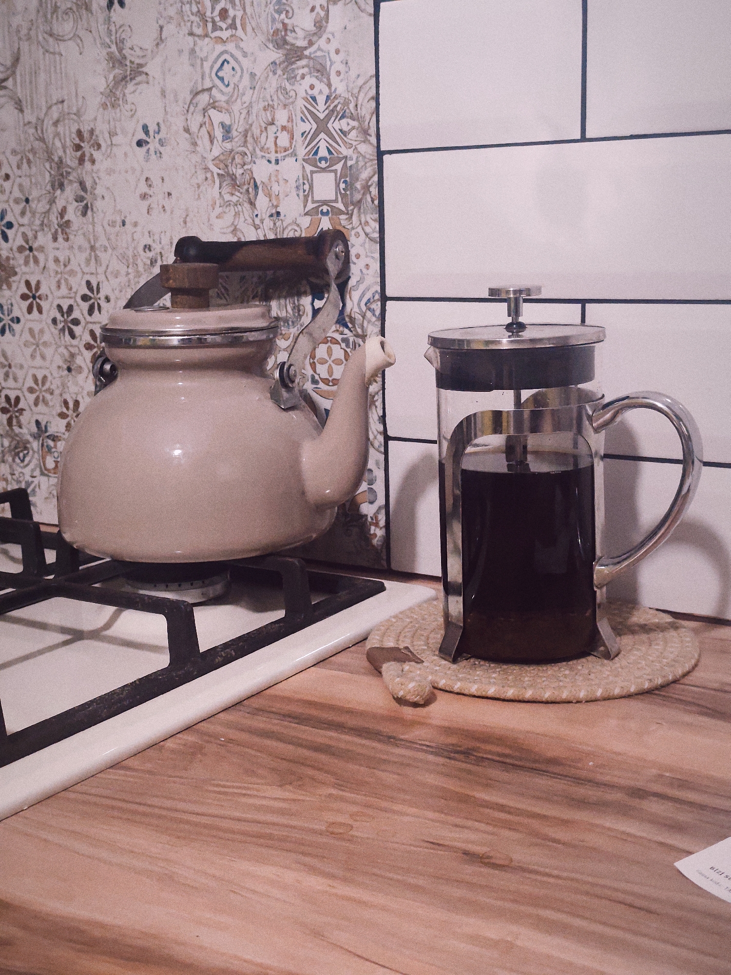 In a domestic kitchen, a beige kettle sits on a gas stove, its wooden handle at an angle. To the right, a glass French press filled with dark liquid rests on a woven placemat on a wooden countertop. The background features a tiled backsplash with a floral pattern on the left and white subway tiles on the right. The scene is lit with artificial light, casting subtle shadows. The location is identified as Sabunchu Raion, Azerbaijan.