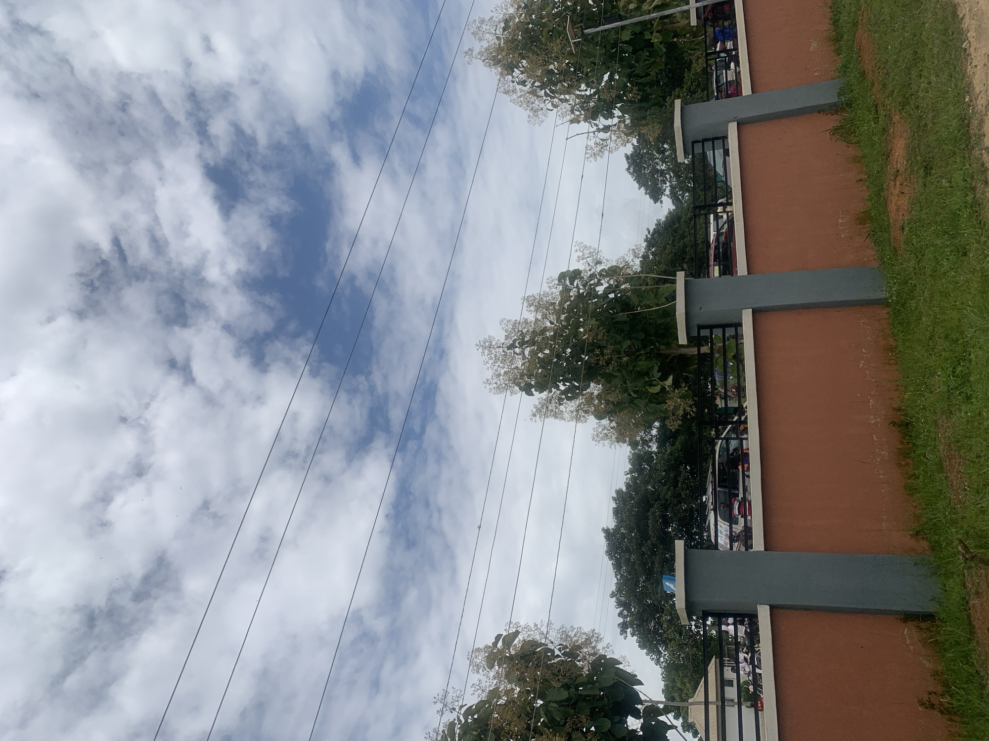 The image shows a cloudy sky with several power lines crossing horizontally. In the foreground, there is a low, reddish-brown wall with grey pillars. Behind the wall, there are trees, some with light-colored blossoms, and parked vehicles, including vans and cars. People can be seen near the vehicles. The scene appears to be an outdoor area in Duk, Nigeria.
