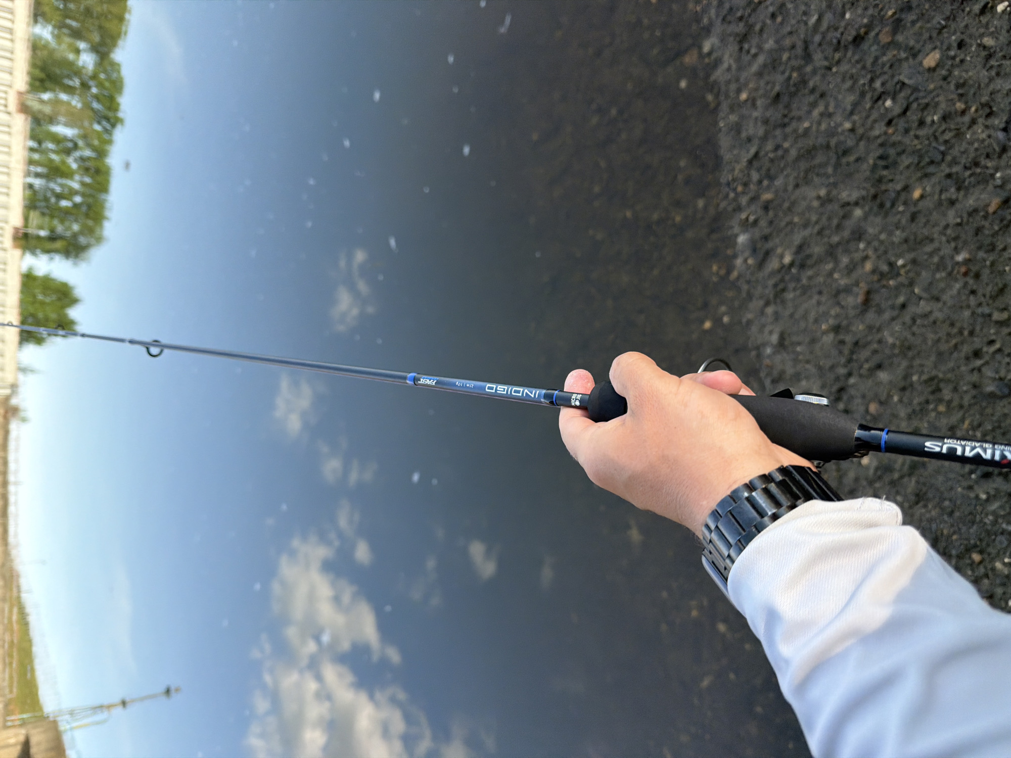 A person is holding a fishing rod in front of a body of water. The rod, a "KIMUS RISING GLADIATOR INDIGO" model, extends horizontally from the right of the frame to the left, with the hand gripping the rod's handle. The person wears a silver and black bracelet watch. The body of water reflects the sky and a building, while the ground to the right is paved and dark. The location is Chaikovsky, Russia.