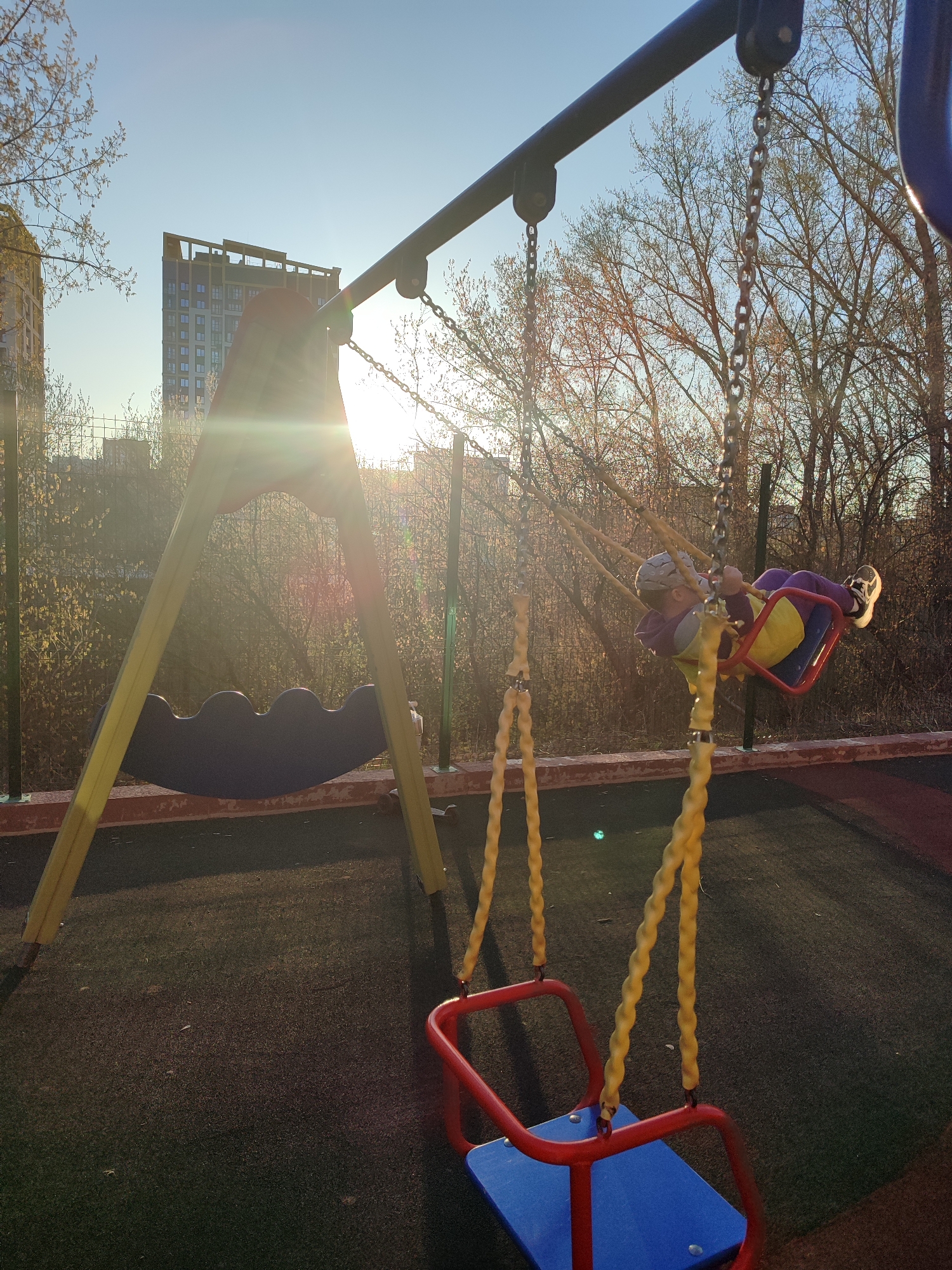 The image depicts a playground scene in Izhevsk, Russia. A child is swinging on a swing set. There is another swing set nearby, with a rocking chair. The playground is covered in a dark, rubberized material. A fence separates the playground from a line of trees and bushes. In the background, a tall building is visible under a clear sky. Sunlight is shining brightly, partially obscuring the view.