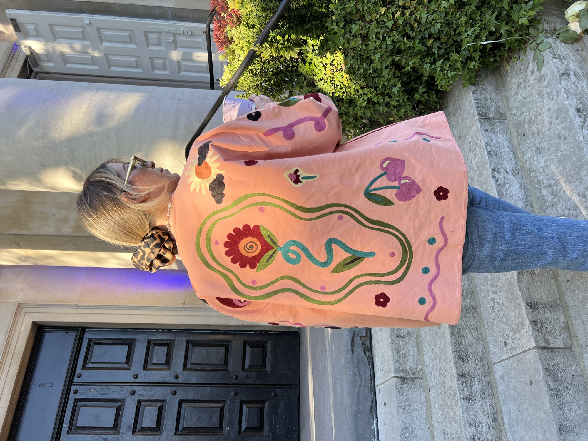 A person is depicted outdoors, seen in profile from their right side, against an architectural background. The individual, appearing to be a woman with blonde hair tied back with a leopard print scrunchie, wears gold-toned sunglasses and a distinctive salmon-pink, long-sleeved jacket. This jacket is intricately embroidered with various colorful motifs, including large stylized flowers (one prominent red and orange with a spiral center), green and teal wavy lines, small purple dots, red cherries, and a sun-like design with dark cloud shapes. She is also wearing blue jeans and holds a white cup, on which the word "TIMELESS" is faintly visible.
The scene takes place on a sunny day, indicated by clear shadows cast across the stone steps and a white door in the background. The time of day appears to be late morning or afternoon. The setting features grey stone steps leading up, with a prominent black panelled wooden door at ground level and part of a white panelled door visible above it. Lush green foliage and bushes are present in the upper right portion of the image, alongside a black metal railing. In one detail, a white rose is seen lying on the stone steps. The overall impression is one of casual style in an urban or residential outdoor environment, possibly in London.