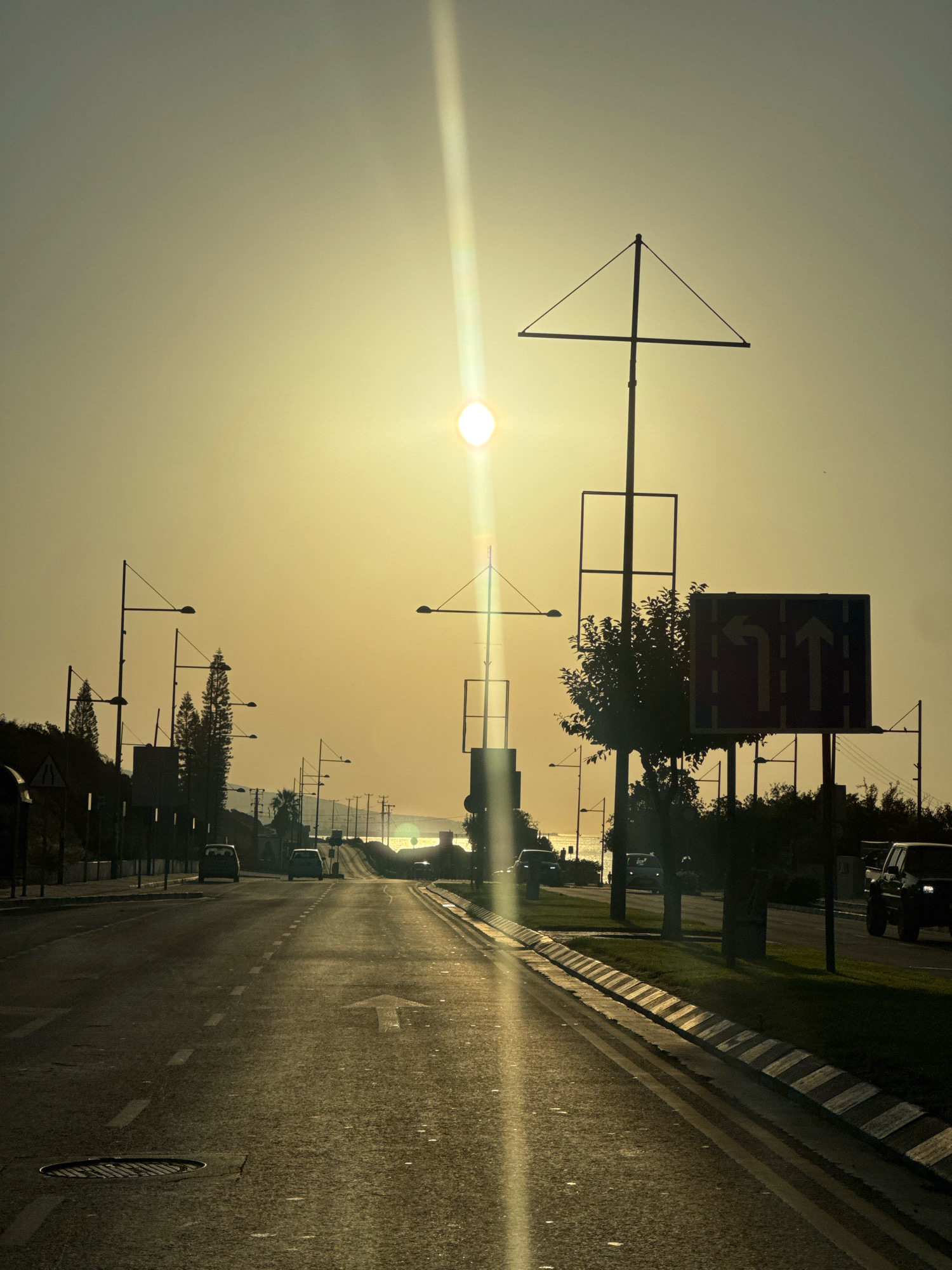 The image depicts a road in the Agios Tychona Community, Cyprus, during a sunset. The sun is visible in the sky, casting a warm glow and creating a lens flare that streaks down the center of the image. The road is asphalt and shows tire marks and painted lane dividers. Several cars are visible on the road, driving in both directions. Along the sides of the road, there are streetlights, trees, and a body of water in the distance. A traffic sign indicating turning lanes is also present. The overall atmosphere is serene and bathed in the golden light of the setting sun.