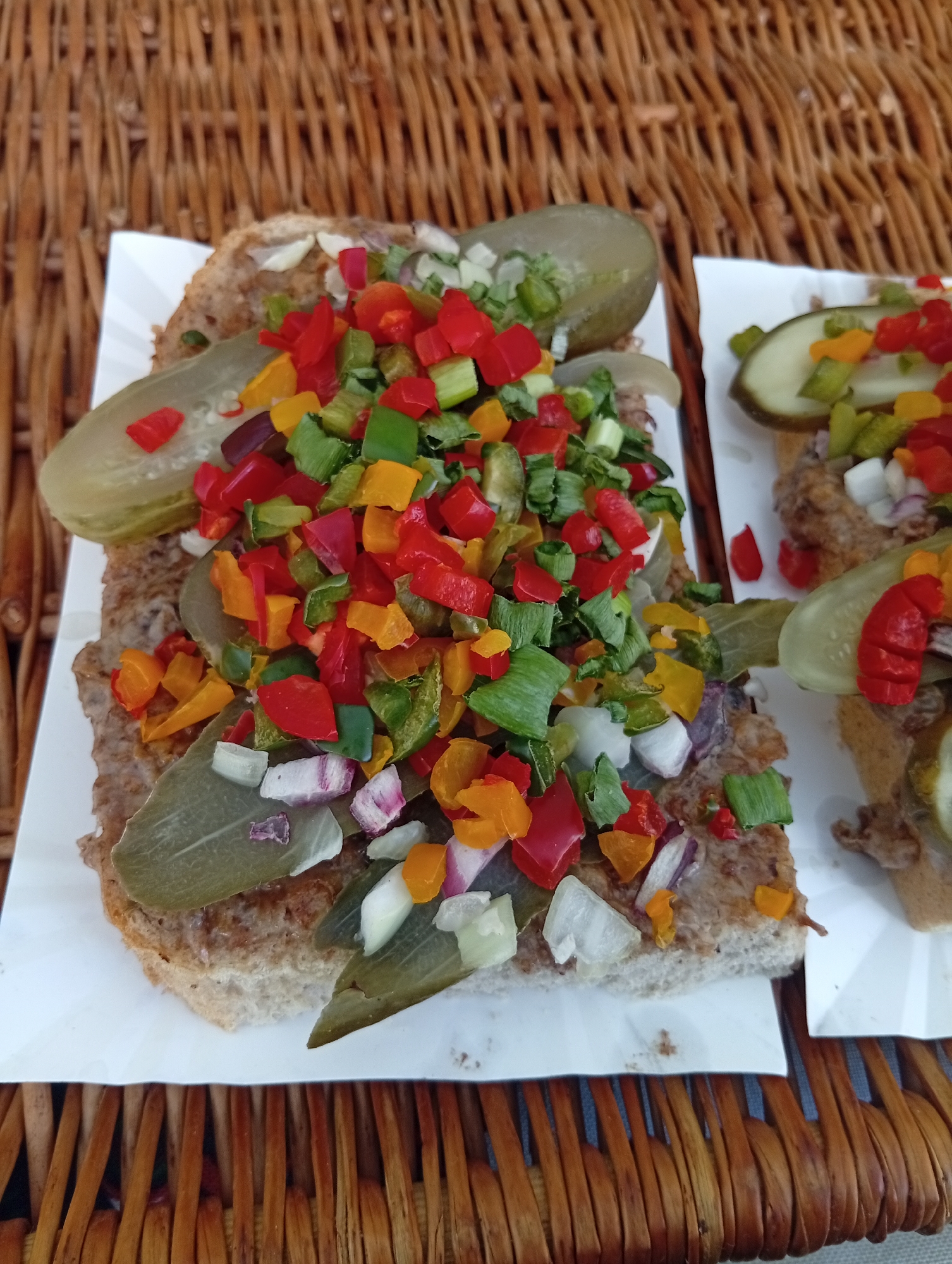 Two open-faced sandwiches, appearing to be a type of toast with a ground meat topping, are presented on white paper plates atop a woven, wicker-like surface. The sandwiches are topped with a generous portion of chopped bell peppers in red, yellow, and green, as well as chopped green onions and what appear to be thinly sliced red onions and pickles. Slices of dill pickles are also visible on the sandwiches, along with some larger, pale green leafy elements, possibly lettuce or a herb. One sandwich is partially visible on the right side of the image, suggesting a market or food stall setting. The background is dominated by the brown, textured weave of the surface the plates are resting on. The overall presentation is rustic and casual. The food appears to be served outdoors, possibly at a street food vendor or an outdoor market in Wrocław, Poland.