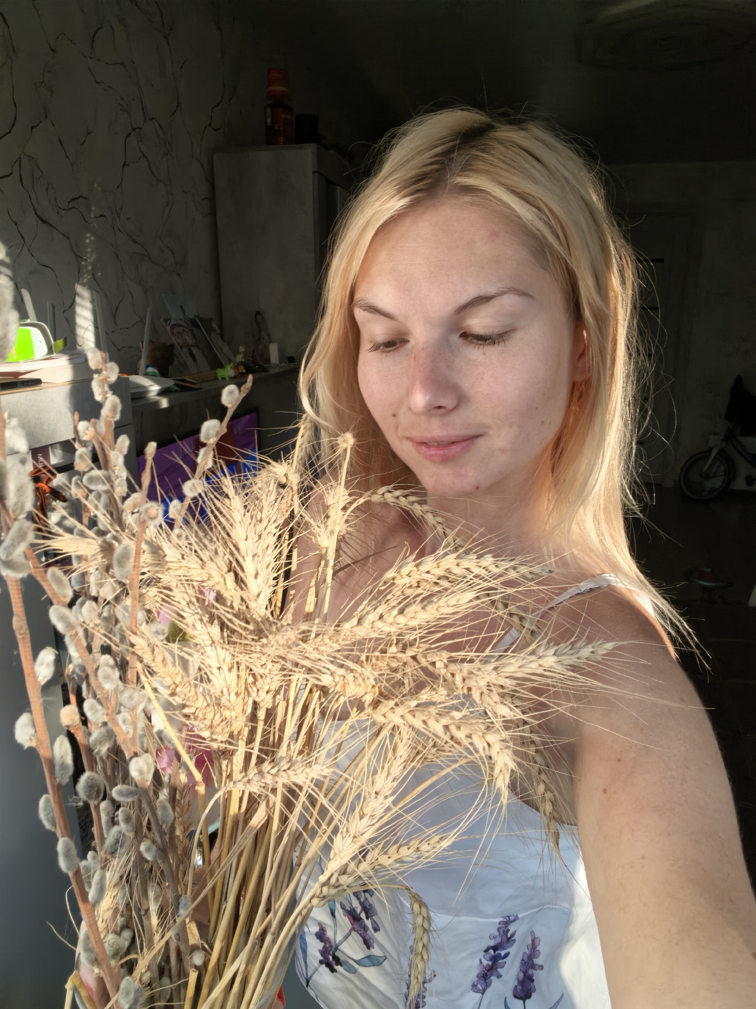 A blonde woman with fair skin and freckles looks down and to her right, holding a bouquet of dried wheat stalks and pussy willow branches. She is wearing a white garment with a floral pattern. The background is dimly lit and appears to be indoors, with a textured wall and shelves holding various items. The lighting suggests it is either early morning or late afternoon, with sunlight streaming in from the left. The overall atmosphere is serene. The specific location in Sterlitamak, Russia cannot be determined from the provided image.