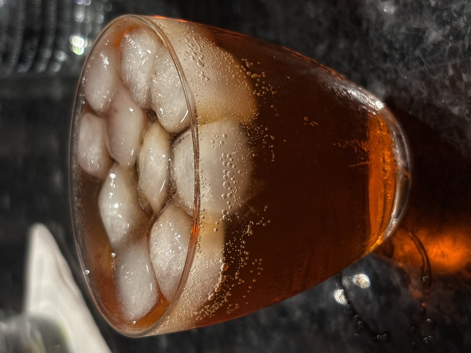 The image displays a close-up, elevated view of a clear, tapered drinking glass containing a reddish-brown, carbonated beverage. Approximately ten to twelve irregularly shaped ice cubes are floating within the liquid, occupying the upper portion of the glass. Numerous small bubbles are visible adhering to the inner surface of the glass and suspended within the liquid, indicating carbonation. The glass rests on a dark, textured surface, possibly granite or a similar material, which reflects subtle light. The background is out of focus, showing dark, indistinct shapes and faint highlights, including the blurred outline of another object, potentially a glass, on a lighter, indistinct surface to the upper left. No discernible features within the image indicate a specific location in Tunbridge Wells, United Kingdom. No people are present.