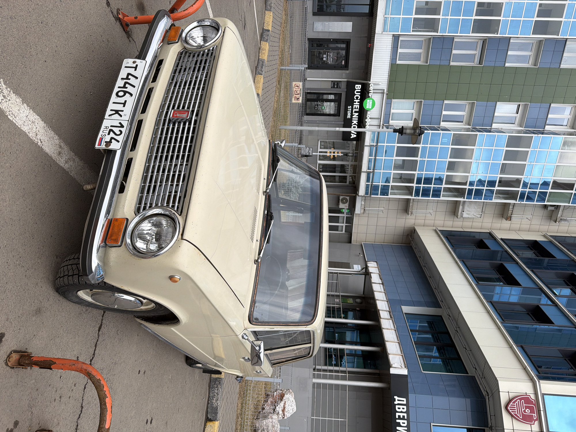 A tan-colored Lada car is parked on a city street. The car has a Russian license plate that reads "T446TK 124 RUS". The backdrop includes a building with a sign in Russian that reads "BUCHELNIKOVA STORE". Further down on another building the word "ДВЕРИ" is visible, and another building is clad in blue and green glass. The location is Krasnoyarsk, Russia.