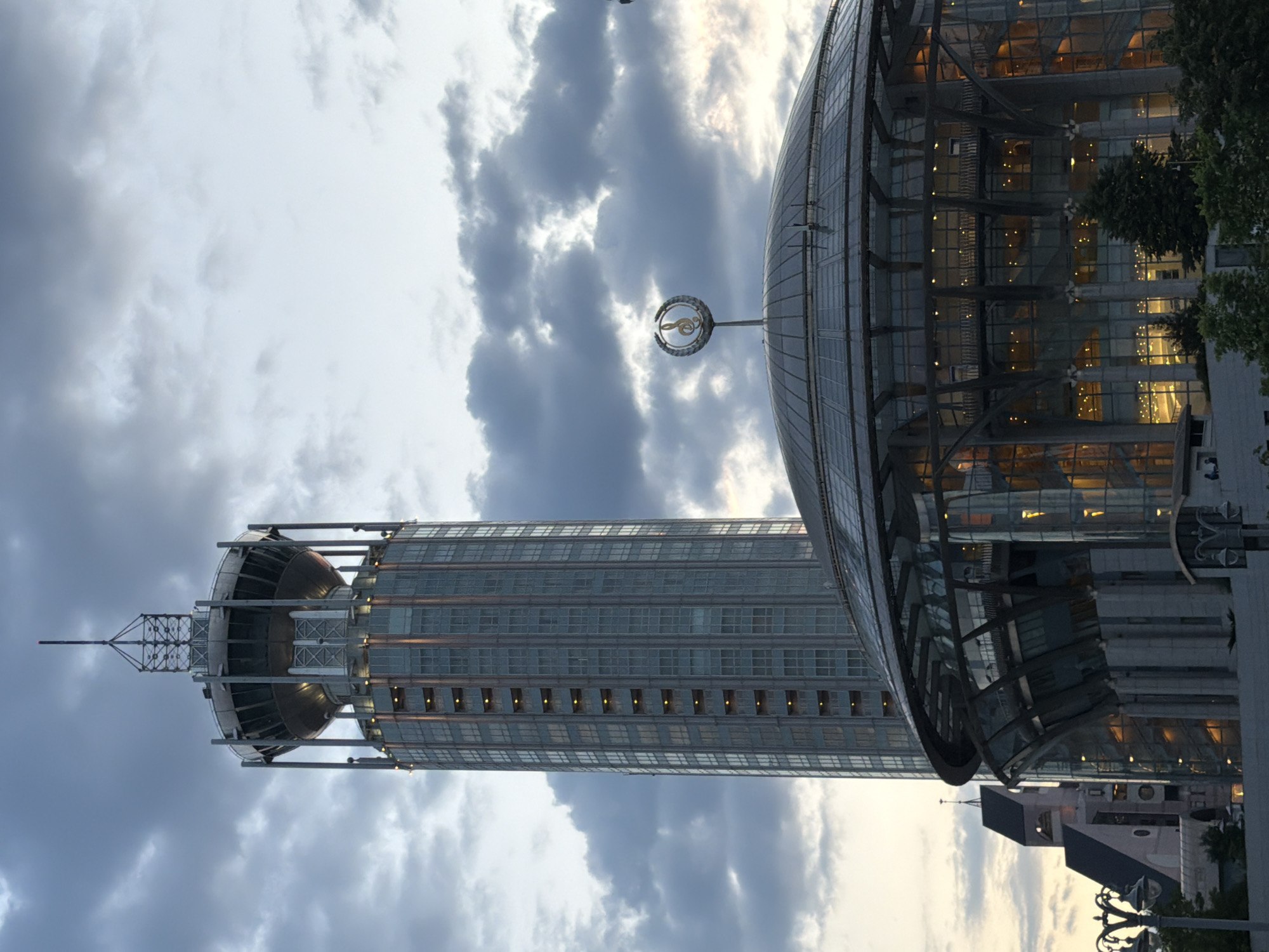 This image captures a modern urban cityscape in Moscow, Russia, under a partly cloudy sky, likely during late afternoon or early evening. The lighting suggests a "golden hour" aesthetic, with warm tones reflecting on glass surfaces and illuminating interiors.
The scene features two prominent architectural landmarks. On the left, a tall, slender skyscraper with a cylindrical body and a pointed spire at its top dominates the frame. Its facade is characterized by numerous rectangular windows, many of which glow with a warm, interior light. This building is identifiable as the Swissôtel Krasnye Kholmy.
To the right, a large, contemporary building with an expansive glass-domed roof is visible. Rising from the center of its dome is a distinct golden emblem, shaped like a stylized musical note or treble clef encircled by a crescent. This emblem is characteristic of the Moscow International House of Music. The building's glass exterior reflects the ambient light and reveals illuminated interiors.
Scattered clouds are present in the sky, with brighter patches indicating the sun's position. Hints of green foliage from trees are visible on the right side, and parts of other urban structures and a streetlight can be seen at the bottom right, grounding the scene in a city environment. No discernible text is visible in the image.
