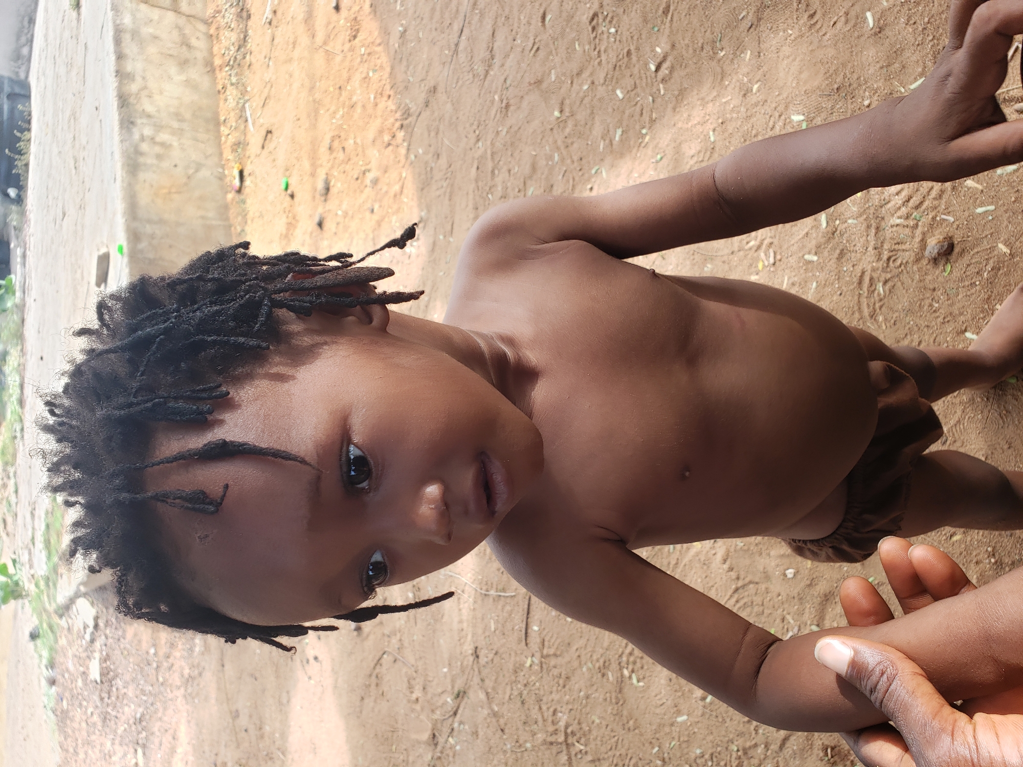 A full-body, portrait-oriented shot captures a young child outdoors, gazing directly at the viewer. The child, likely a toddler, has dark skin, large dark eyes, and short dreadlocks that frame their face. They are bare-chested, wearing only a pair of dark brown, ruffled diaper cover or small shorts.
The setting is an open, sunlit area with light brown, sandy or dirt ground. Small green leaves and scattered debris are visible on the ground. In the background, to the left, a light-colored concrete structure or wall stands, with a few faint green objects visible near its base.
An adult's dark-skinned hand enters the frame from the lower right, gently holding the child's right arm, indicating support or guidance. The child's expression is calm and direct, showing a natural curiosity. The bright illumination suggests it is daytime, likely sunny. The overall scene conveys a simple, natural outdoor environment, consistent with the Adankolo, Nigeria context. There is no visible text.