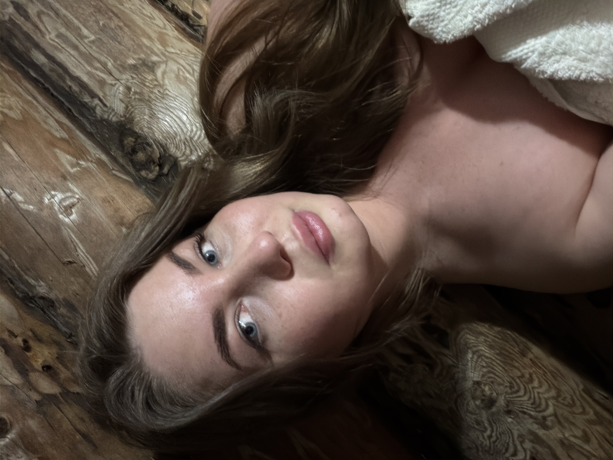A light-skinned woman with brown hair and blue eyes lies on a wooden surface. Her head is tilted towards the viewer. A white towel is partially visible near her shoulder. The location is Уральское, Russia.