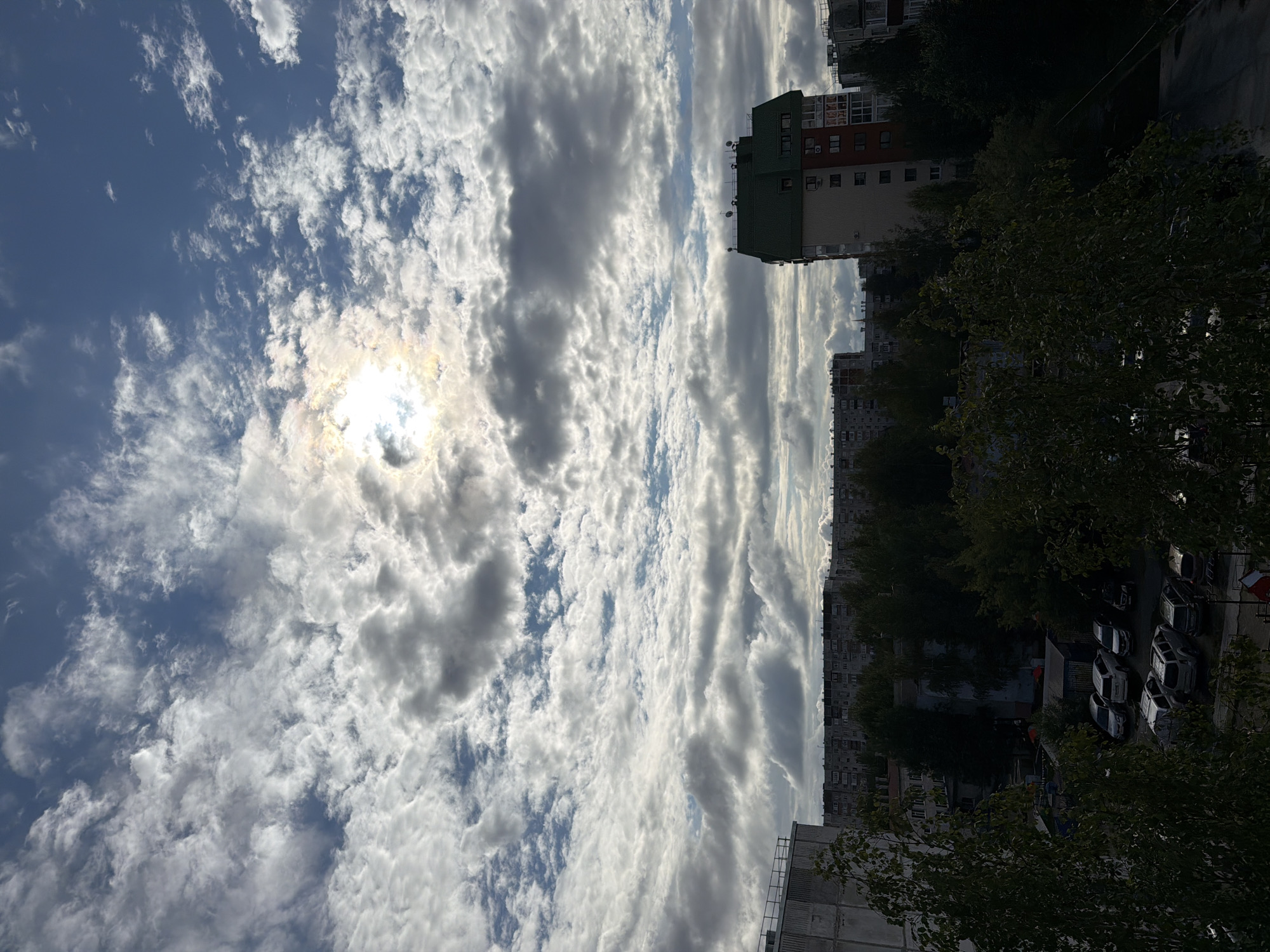 The image depicts a cloudy sky over a residential area in Surgut, Russia. The sun is partially obscured by clouds, creating a bright halo. In the foreground, trees with dark green foliage frame the scene. Beyond the trees, several apartment buildings are visible, including one with a distinctive green roof. A parking lot with several cars is situated in front of the buildings. The overall impression is that of a typical urban landscape on a cloudy day in Surgut.