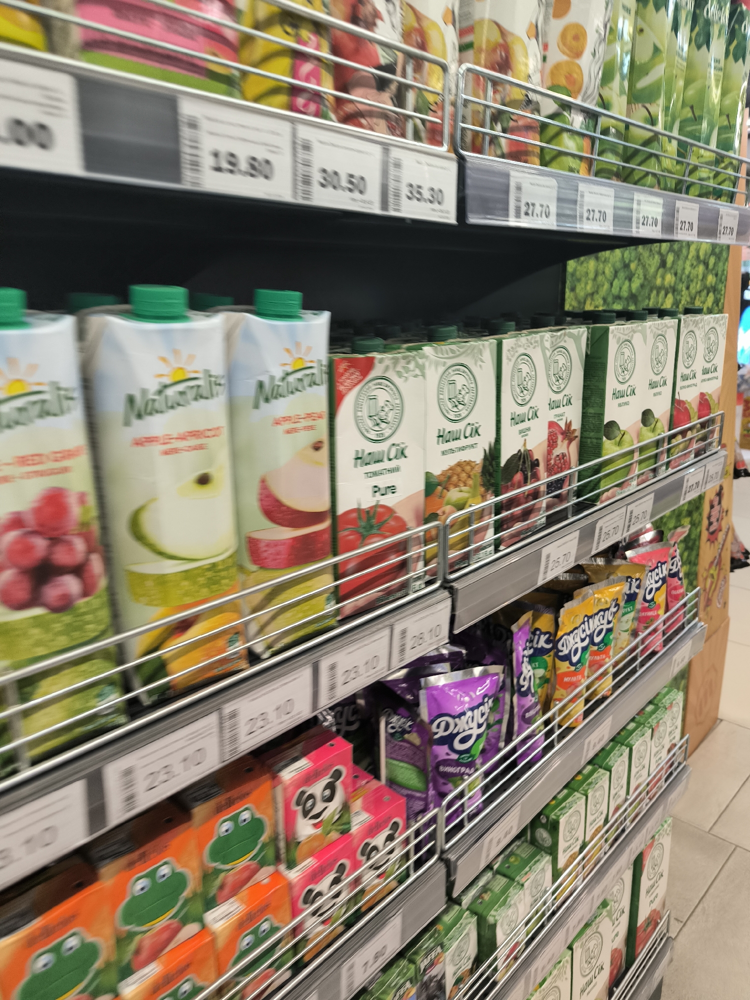 The image shows a retail display of juice cartons and pouches on multiple shelves in a supermarket. The products are arranged in rows, with various brands and flavors visible. Prices are displayed on labels below each shelf. The shelves appear to be made of metal, and the overall environment suggests a well-stocked grocery store. The products include cartons with images of fruits such as grapes, apples, pears, and pomegranates, as well as some cartoon characters. There are also pouches with brightly colored designs. The lighting is typical of a retail environment, with overhead lights illuminating the products. The scene is viewed from a slightly elevated angle, looking down at the shelves.