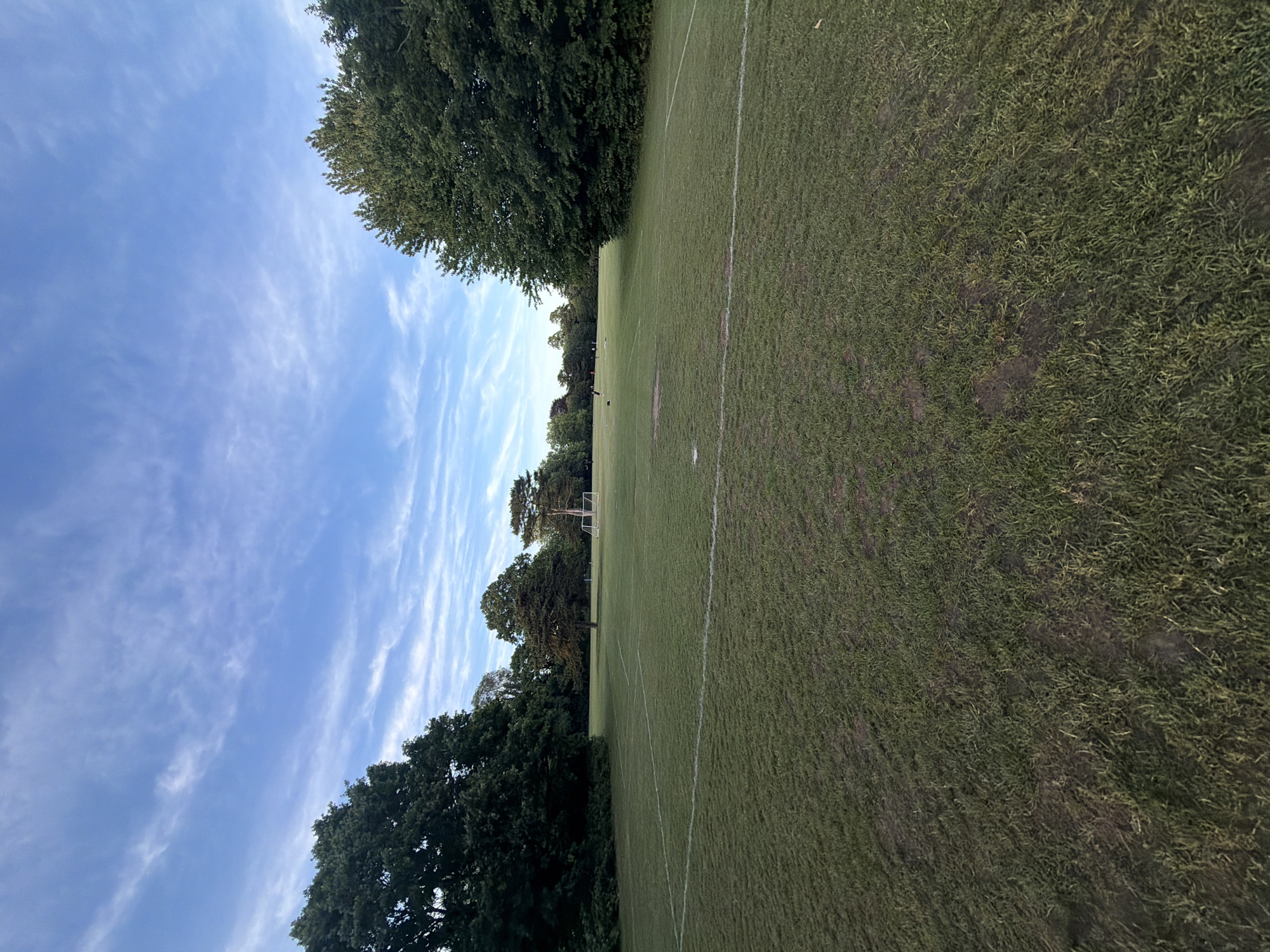 This image depicts a large, open sports field in Maidstone, United Kingdom, under a clear blue sky interspersed with wispy white clouds. The foreground and midground consist of green grass, exhibiting areas of wear and bare earth. Faint white lines are visible on the grass, delineating sections of a sports pitch. A white football goal is positioned in the middle distance, slightly to the right of the central axis. The field is bordered by dense deciduous trees on both the left and right sides, as well as in the background. In the far distance, several small, indistinct human figures are visible on the field, gathered in a group, suggesting recreational activity.