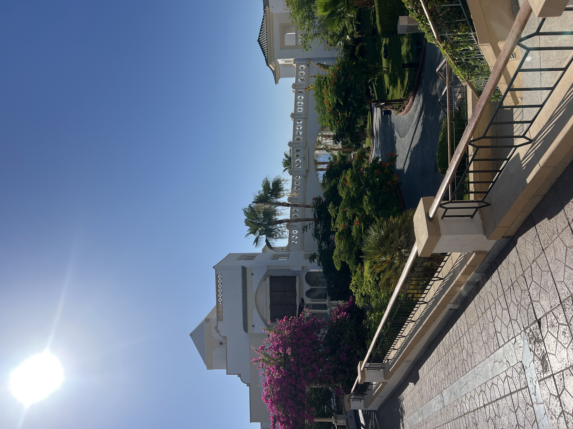 The image depicts a bright, sunny day in Sharm El Sheikh, Egypt, at what appears to be a resort. The foreground features a textured stone pathway with a decorative metal railing. Beyond this, a multi-level white building with archways and ornate windows is visible, flanked by lush green foliage and a vibrant pink bougainvillea bush. Further in the background, a white bridge-like structure connects two parts of the building, with palm trees and other tropical vegetation surrounding it. The sky is a clear, bright blue, with the sun prominently visible in the upper left quadrant, casting long shadows. The overall scene conveys a sense of resort luxury and a warm, Mediterranean climate. There are no people visible in the image.