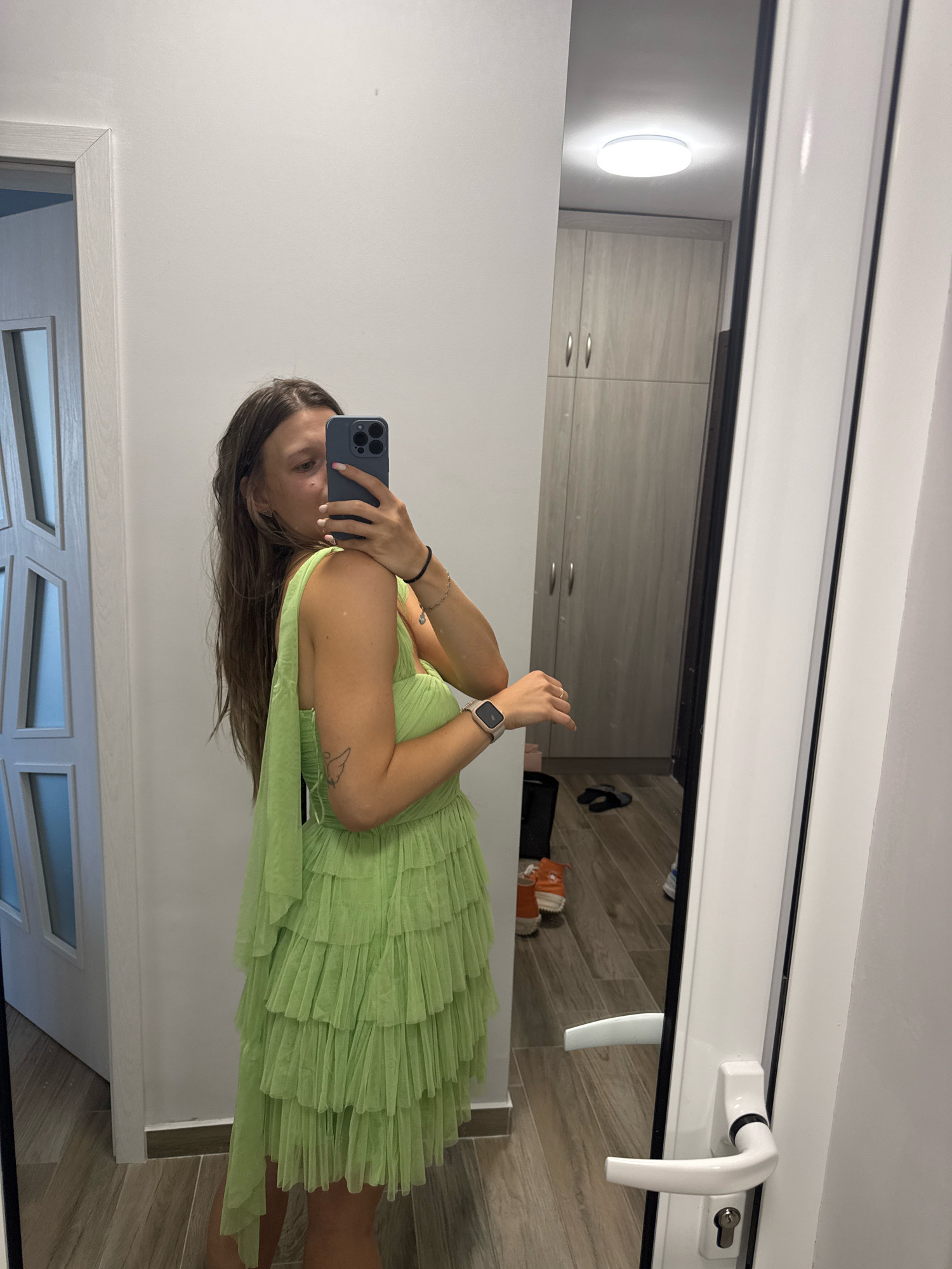 A young woman is taking a selfie in a room that appears to be a bedroom or dressing room. She is wearing a light green, tiered, tulle dress and is holding a dark blue smartphone in her right hand to take the photo. The room has light wood flooring and grey wooden cabinets. A large mirror is positioned to her right, reflecting the scene. Through the mirror, a white door with a glass panel is visible on the left, and what looks like an external door handle can be seen on the right. The image does not provide any specific visual cues to determine the location within the city of Sofia, Bulgaria.