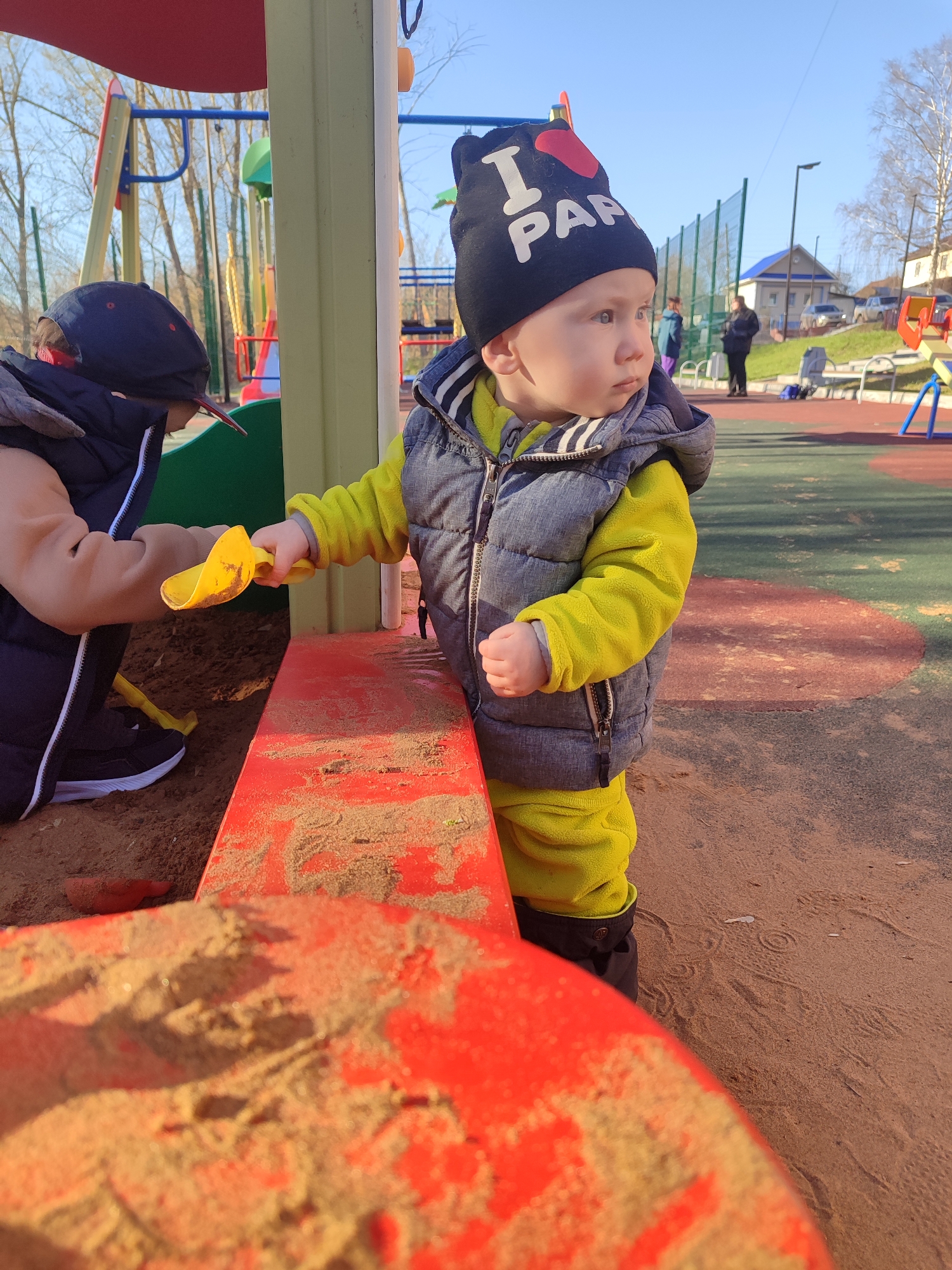 Two young children are at a playground in Izhevsk, Russia. One child, wearing a black hat that says "I ❤ PAR," a gray vest, and a bright yellow outfit, stands near a red sandbox. The child is looking to the right, away from the sandbox. The other child, wearing a dark blue cap, is leaning over the sandbox, holding a yellow plastic scoop. Both children appear to be playing with the sand. The playground has a soft, rubbery surface. In the background, there are trees, houses, and other playground equipment. The weather appears sunny and clear.