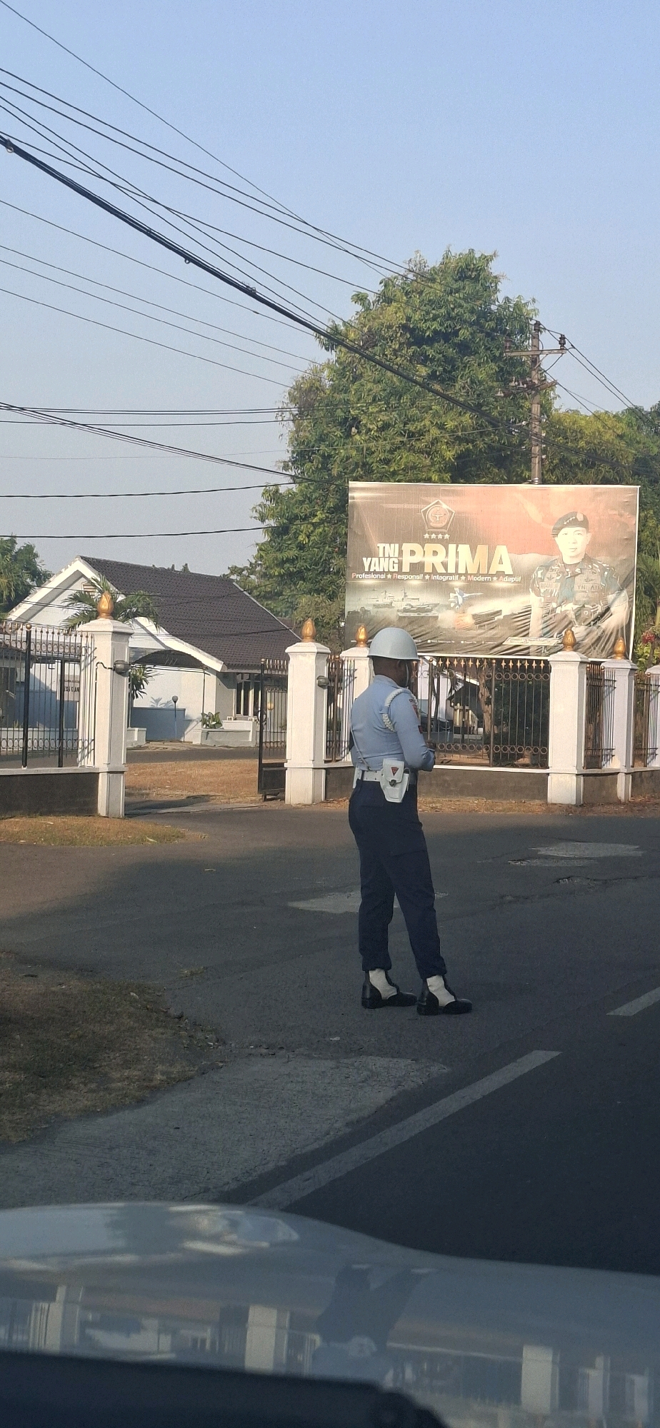 A man in a light blue uniform and dark blue pants stands on an asphalt road, facing away from the camera, in front of a white fence with ornate pillars. He wears a white helmet and a white utility belt with a red and white pouch. Behind the fence is a white house with a dark roof and a large banner displaying the words "TNI YANG PRIMA" and an image of a military officer. The scene is set outdoors, with trees and power lines visible in the background. The location is identified as Colomadu, Indonesia, likely near a military facility given the banner. The lighting suggests it is daytime, possibly morning or afternoon.