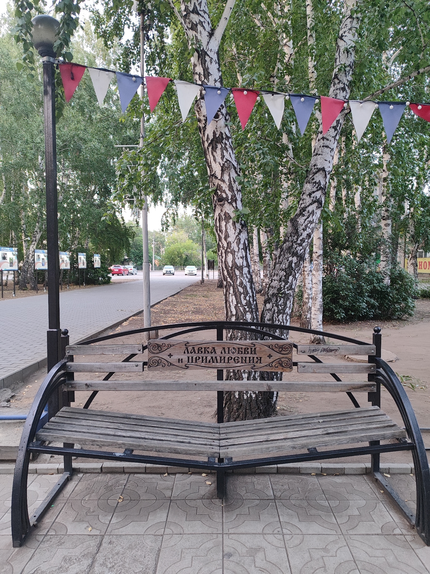 This is a medium-shot, eye-level, outdoor photograph of a park bench in Slavgorod, Russia. The bench is made of dark metal with a curved frame and wooden slats. A wooden plaque is attached to the backrest of the bench. The plaque has ornate carvings on the sides and text in Cyrillic that reads "ЛАВКА ЛЮБВИ И ПРИМИРЕНИЯ," which translates to "Bench of Love and Reconciliation." A string of triangular flags in red, white, and blue hangs across the top of the bench, attached to a lamppost on the left and tree branches. The bench is situated on a paved area with a patterned tile design. Behind the bench, there are several birch trees with distinctive white bark and green foliage. Further in the background, a paved walkway lines a street with cars and informational display boards. The overall scene suggests a public park or promenade within the city.