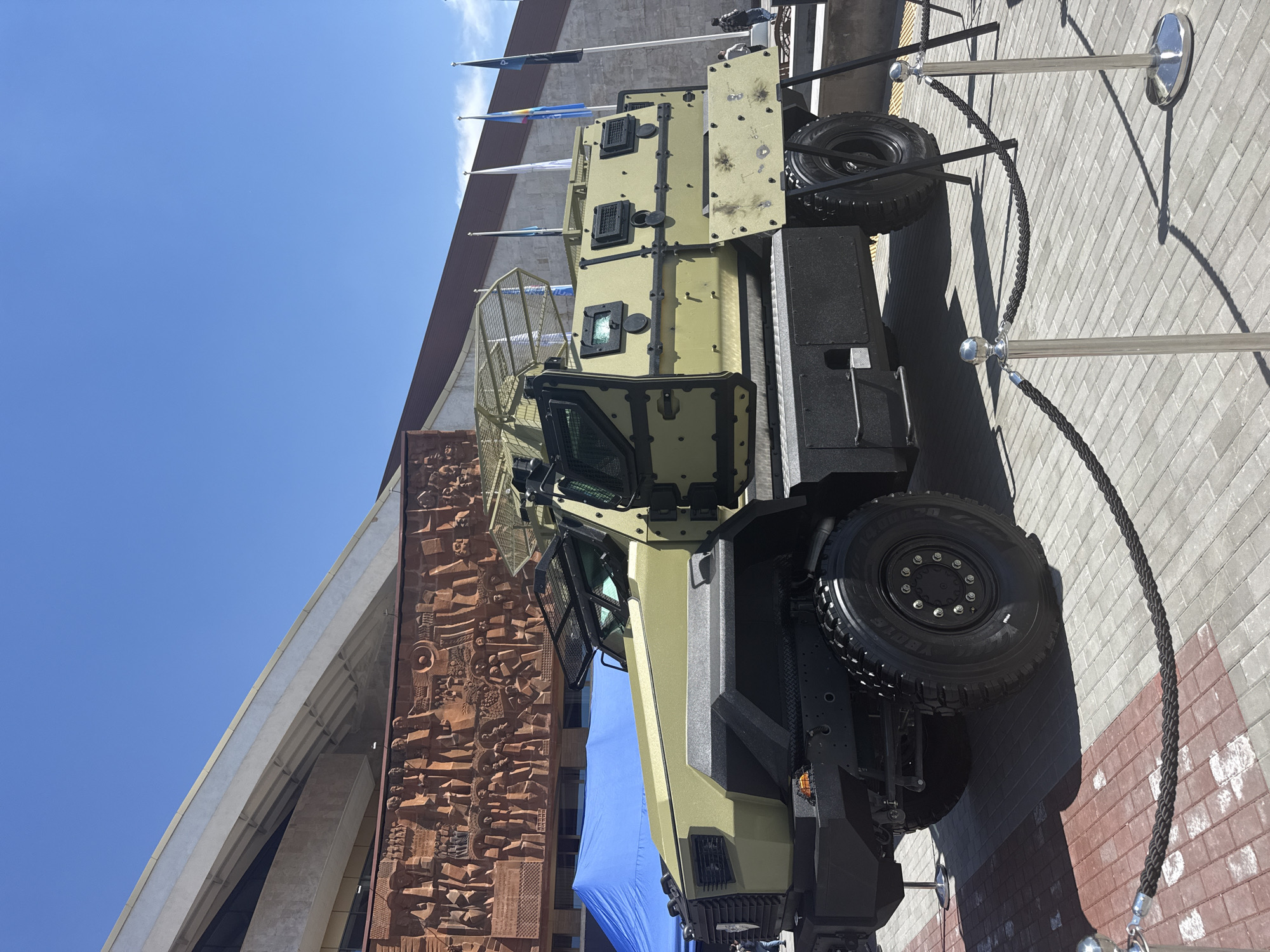 The image, rotated 90 degrees clockwise, displays a large, olive green armored military vehicle, possibly an MRAP, exhibited outdoors under a clear blue sky. The vehicle is parked on a paved surface of gray and reddish-brown interlocking bricks, demarcated by a barrier of chrome stanchions linked by a black braided rope.
The armored vehicle features robust construction with thick plating, large off-road tires (visible text "YOKOHAMA GEOLANDAR G003"), and protective grates over its windows and other openings. A spare tire is mounted on the rear of the vehicle. The armor panels show some scuff marks or minor damage.
In the background, a substantial building is partially visible. It features a large, ornate reddish-brown sculpted relief or mural on one section, while another part displays modern concrete architectural elements. Several flags, some with blue and white colors, are flown from poles near the building. The scene is brightly lit by the sun, casting distinct shadows and suggesting a clear, sunny day, likely mid-day or early afternoon. No individuals are visible in the image. The setup implies the vehicle is on public display, perhaps at an exhibition or museum.