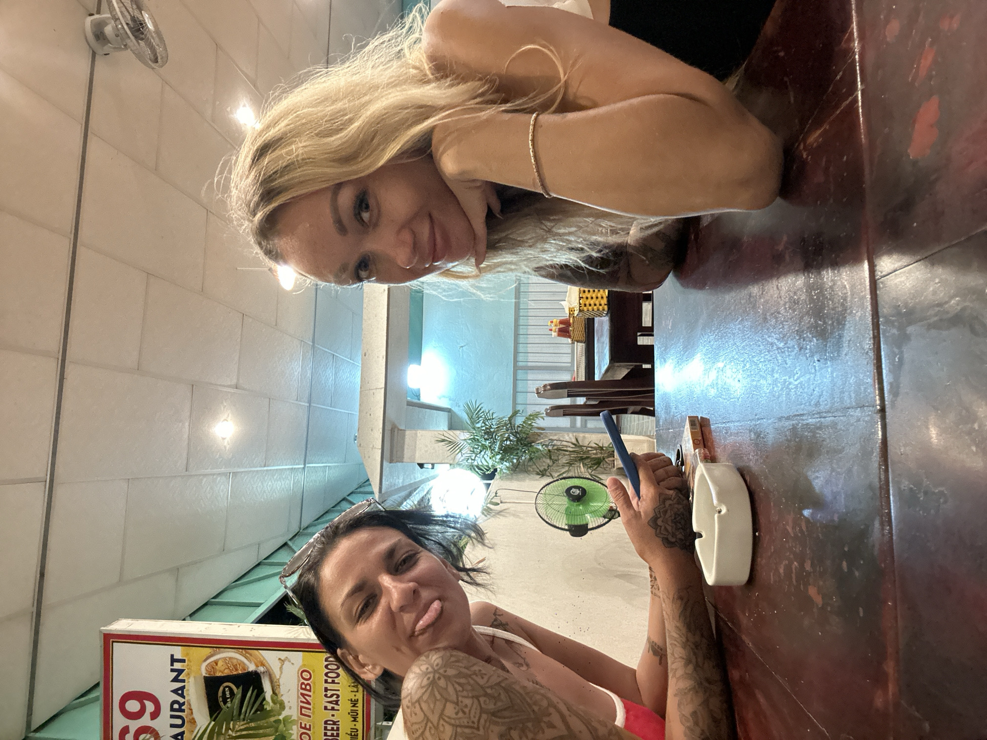 This media file captures a casual selfie taken by two women, likely friends, at an outdoor or semi-outdoor restaurant in Phan Thiết, Vietnam. The image is oriented vertically.
In the foreground, one woman with dark hair, wearing a white tank top and adorned with extensive arm tattoos, is leaning forward and sticking her tongue out playfully towards the camera. She is wearing sunglasses on top of her head. Next to her, a white ashtray is visible on a dark wooden table, and she is holding a blue object, possibly a phone.
In the background, another woman with long blonde hair is lying down on her forearms, also looking towards the camera with a smile. She is wearing a light-colored top. The setting appears to be a casual dining establishment, with a rustic wooden table dominating the right side of the frame. A small fan is mounted on the wall behind the women, and there is a sign for a restaurant with the number "69" prominently displayed, along with text in Vietnamese and English that includes "AURANT," "BEER - FAST FOOD," and place names like "MŨI NÉ." The lighting suggests it could be evening or late afternoon, with artificial lights providing illumination. The overall mood is relaxed and friendly, indicative of a shared meal or outing.