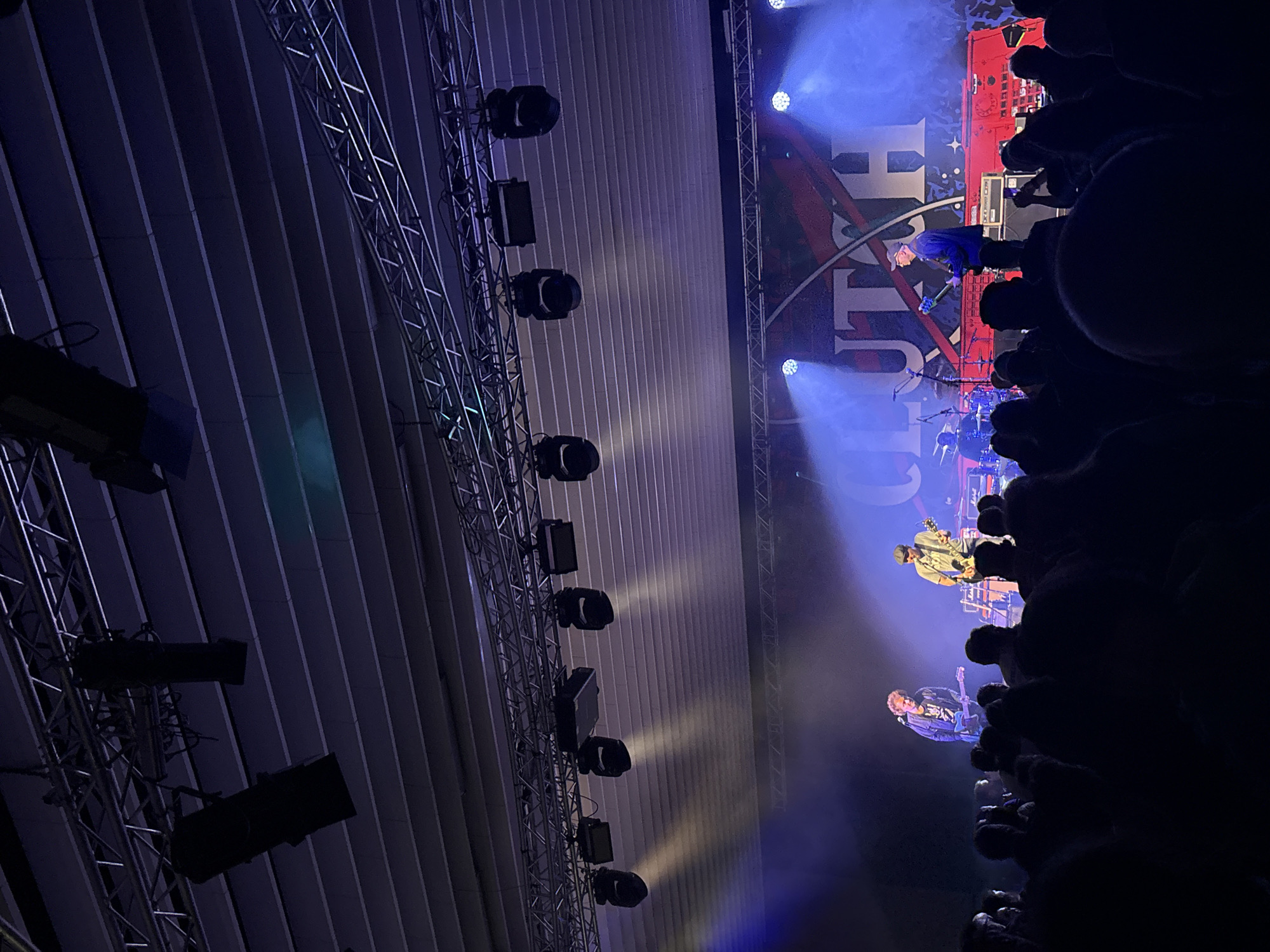 The image depicts a live music performance at an indoor venue. Several musicians are on stage under blue stage lights, performing for a crowd. The stage has a backdrop with the word "CLUTCH" visible. Overhead, there is a lighting truss with multiple lighting fixtures. The building's ceiling consists of parallel linear panels. The location appears to be in Sofia, Bulgaria, based on the band's tour schedule and the event locations they perform at. The crowd is mostly silhouetted in the foreground, with focus on the stage and performers.