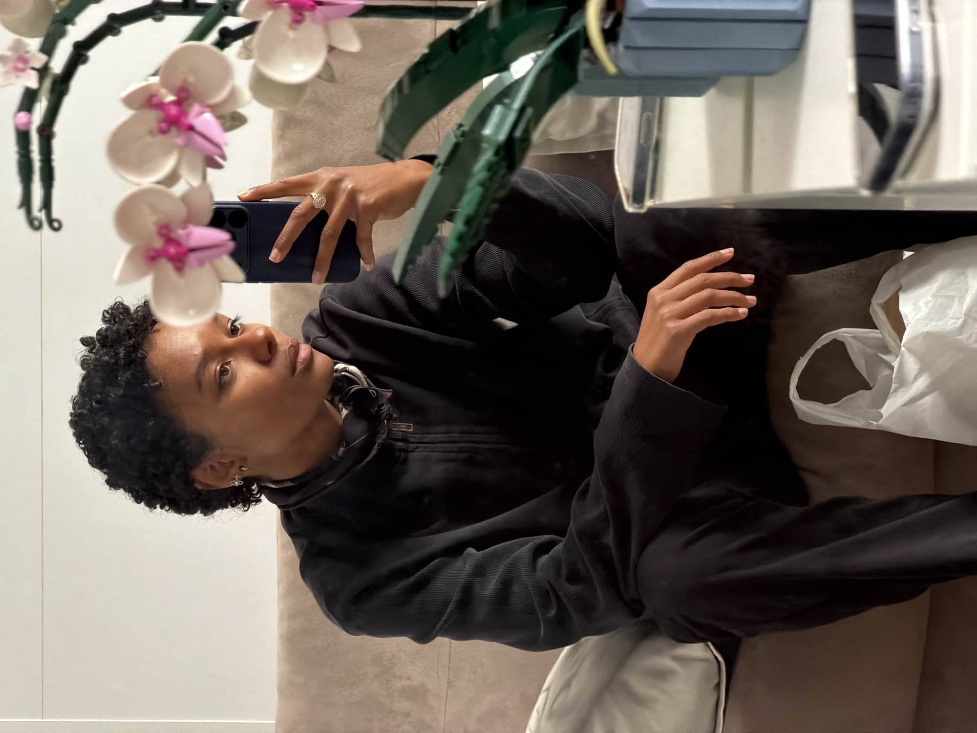 A person with dark curly hair is taking a selfie. They are holding a blue smartphone in their left hand, on which a gold ring is visible. The person is wearing a black zip-up jacket. The background includes a white wall, a beige couch, a white plastic bag, and an artificial plant with pink and white flowers. There is also a glass surface, likely a table or a mirror, reflecting light. The location is not discernible as Paris, France from the given image details.