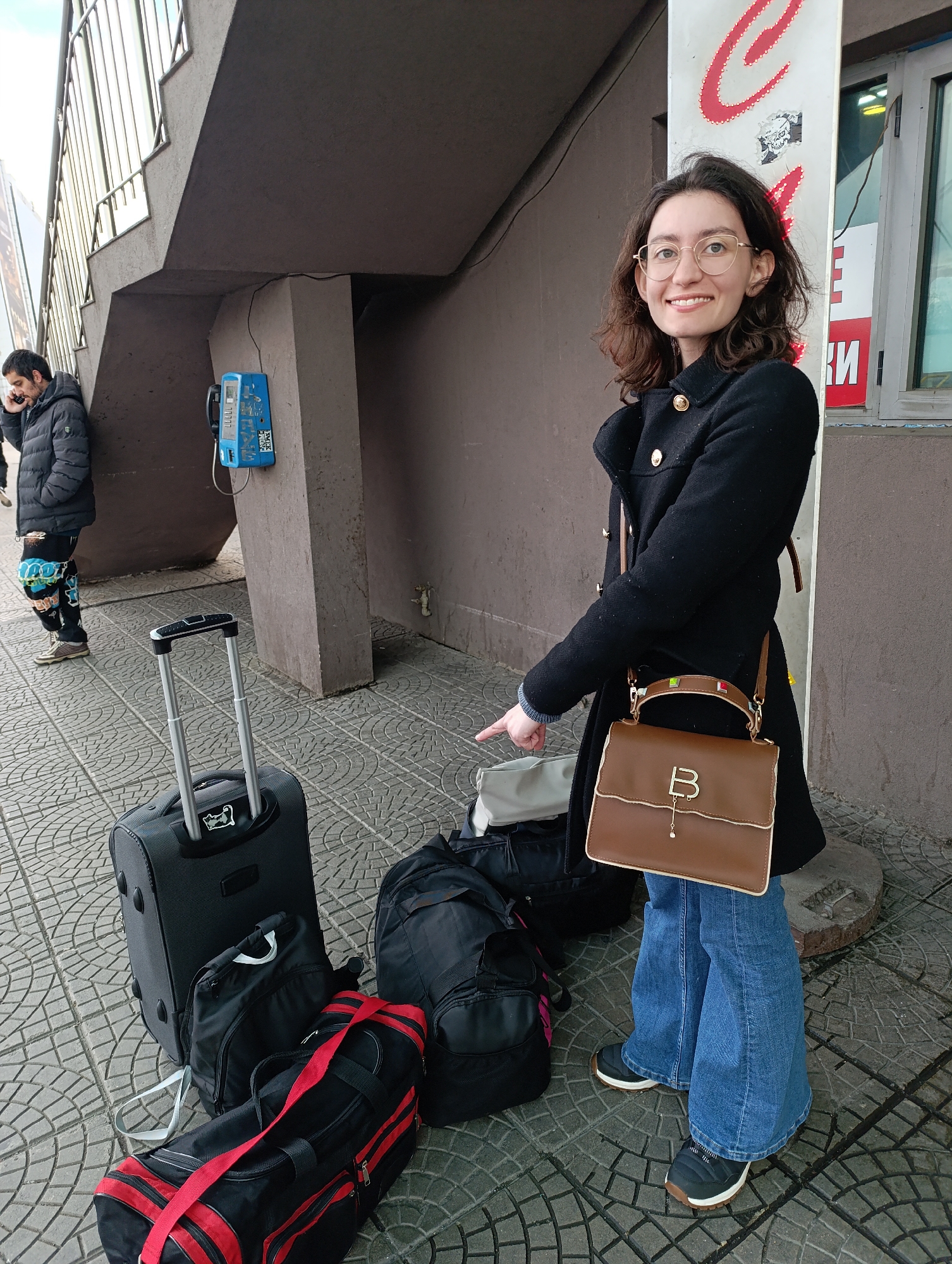 The image shows a woman with glasses and a black coat standing on a tiled sidewalk next to several pieces of luggage: a rolling suitcase, a black duffel bag, and a red and black sports bag. She is pointing towards the luggage with her right hand. She is also wearing blue jeans and black and white sneakers, and a brown purse with the letter "B" on it. Behind her is a concrete wall with a column and a sign on it. To the left of the woman, a man stands next to a vintage blue public telephone. This location is identifiable by the tiled sidewalk, the design of the overpass, the specific look of the public telephone, the design of the building, and the design of the signs as being in Sofia, Bulgaria.