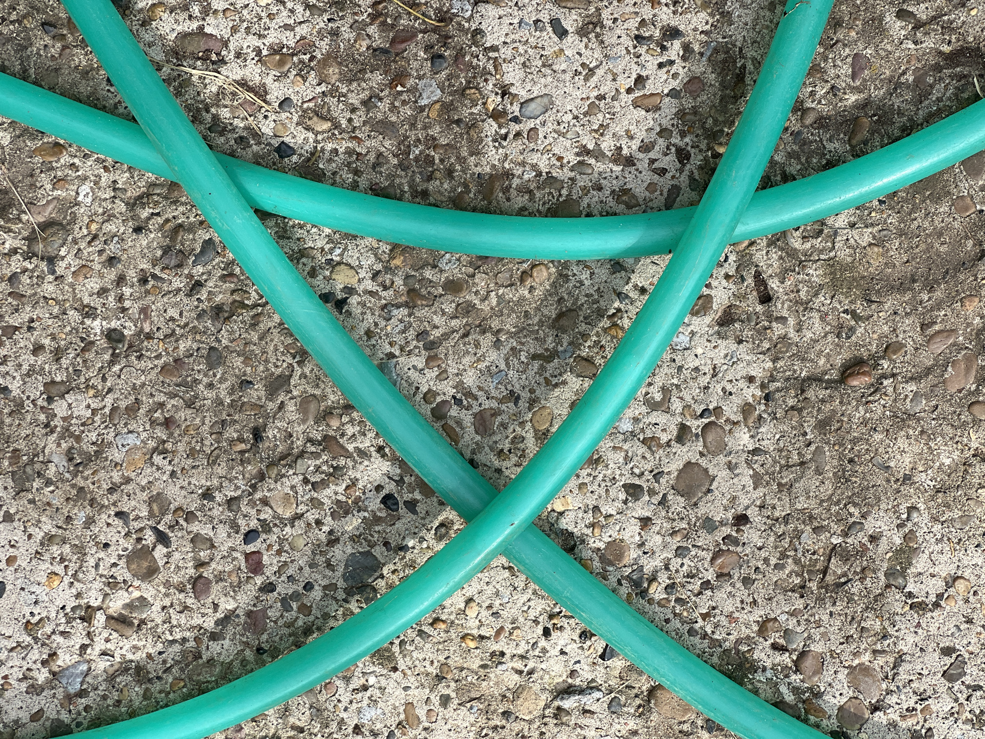 A bright green garden hose lies coiled and crossed over a concrete surface speckled with small, rounded pebbles of various earth tones. The concrete appears weathered and slightly uneven. The lighting suggests it is daytime, with shadows subtly highlighting the texture of the hose and the ground. There are no people or animals visible in the image. The scene is a close-up, overhead view of the hose and pavement, emphasizing the contrast in textures and colors. The location within Yakshur-Bod'ya, Russia is noted as an outdoor setting, likely a yard or driveway.