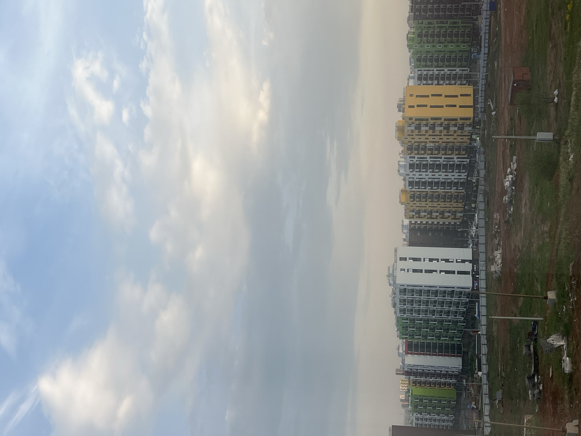 The image, when correctly oriented, presents a wide view of a modern urban landscape in Izhevsk, Russia, dominated by newly constructed residential buildings under a vast, cloudy sky.
On the right side of the frame, a series of multi-story apartment buildings stretches across the horizon. These buildings feature distinct, colorful facades, including bright yellow, clean white, and various shades of green, with some appearing dark grey or black. Their repetitive window patterns and balconies suggest a large-scale housing development.
In the foreground and between the buildings, the ground is a mix of bare brown earth, patches of sparse grass, and scattered construction debris, indicating an ongoing development or recently completed construction site. Utility poles with some attached equipment are visible, running parallel to the buildings.
The left side of the image is filled with the sky, which transitions from a clear, light blue with scattered white, wispy clouds on the far left to a more uniform, hazy light grey or off-white towards the right. This suggests an overcast or partly cloudy day with significant atmospheric haze.
The lighting indicates it is daytime, likely mid-morning or afternoon, with soft, diffused light due to the cloud cover. No people or active events are visible in the scene, conveying a sense of quiet or ongoing development. No discernible text is present in the image.